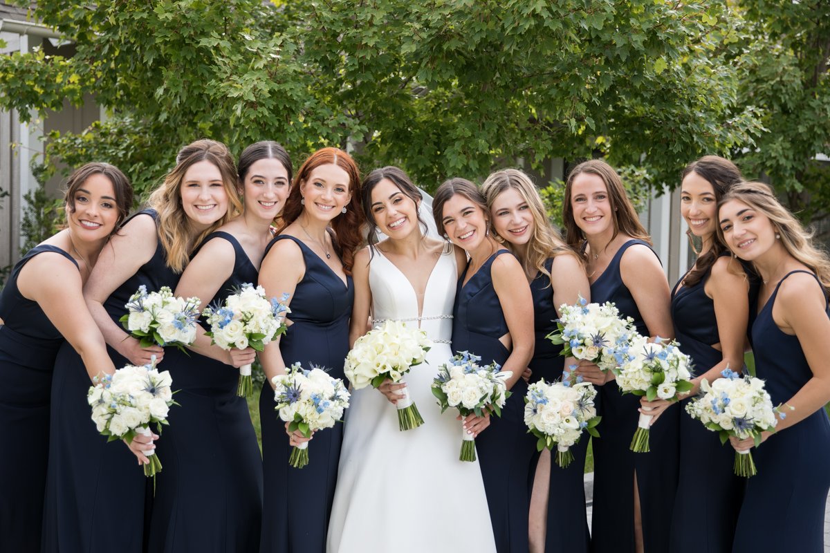 Classic bridal photo with bridesmaids in a line, embracing, smiling with flower bouquets.