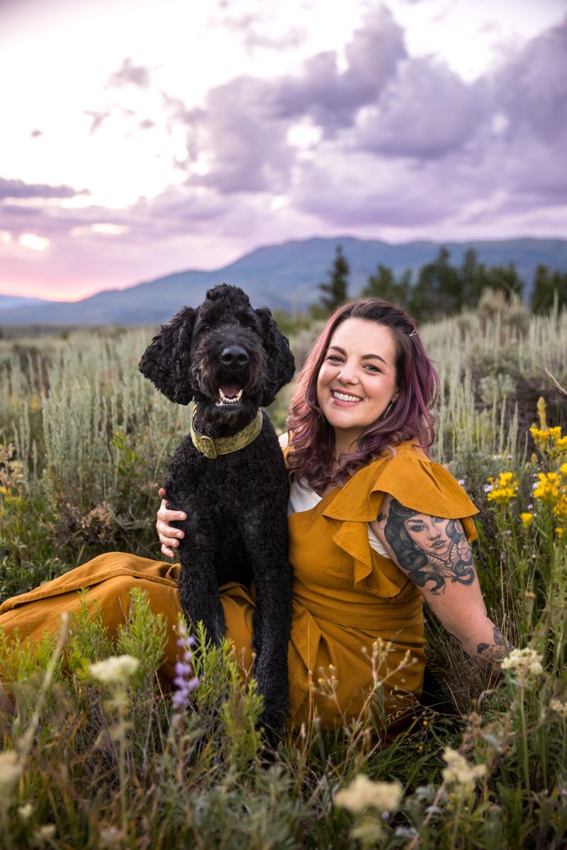 Woman in yellow dress with tattoo sits in a field with wildflowers with a berniedoodle dog on her lap. Pink and purple clouds and mountains are behind her.
