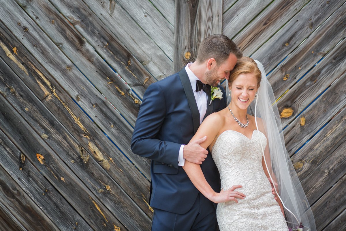 Bride leans into her groom in front of a geometric wooden Wall. They laugh together.