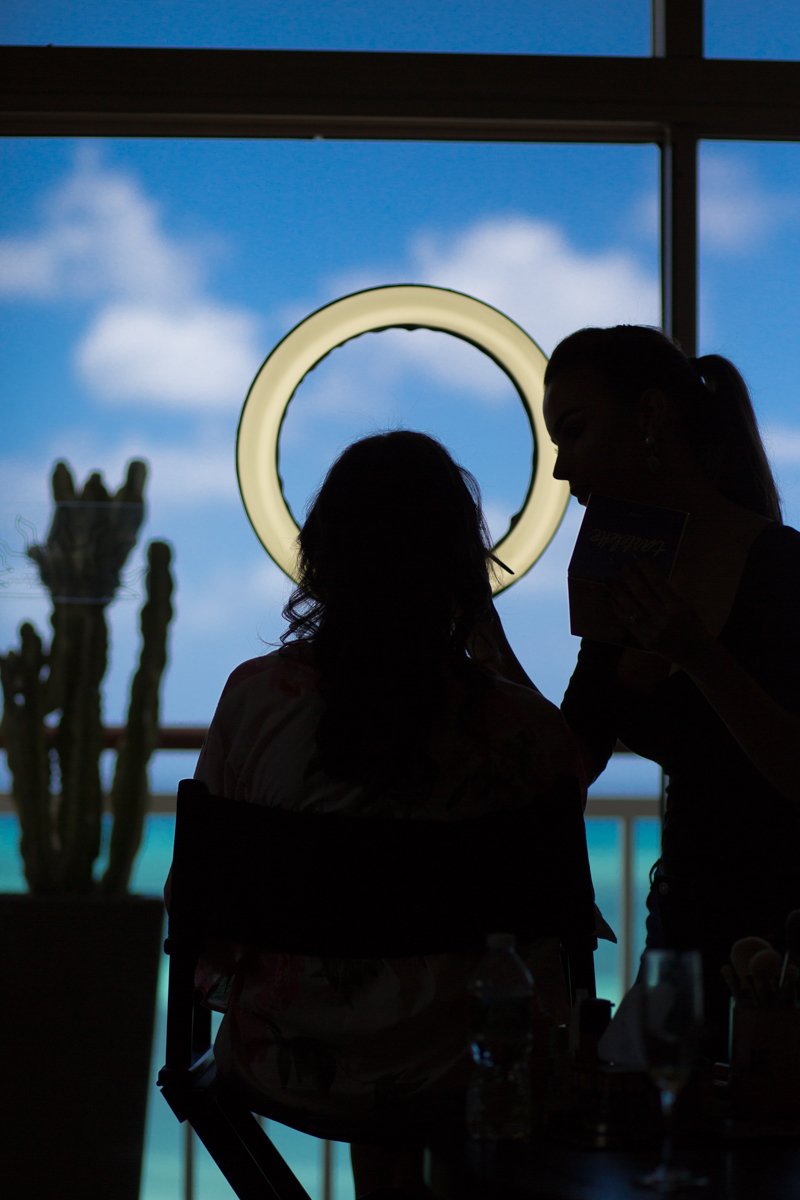 bride getting ready. She sits in a chair as a makeup artist works on her makeup. The shot is silhouetted from behind. A ring light is illuminated. The ocean and blue sky with fluffy white clouds is out the window view.