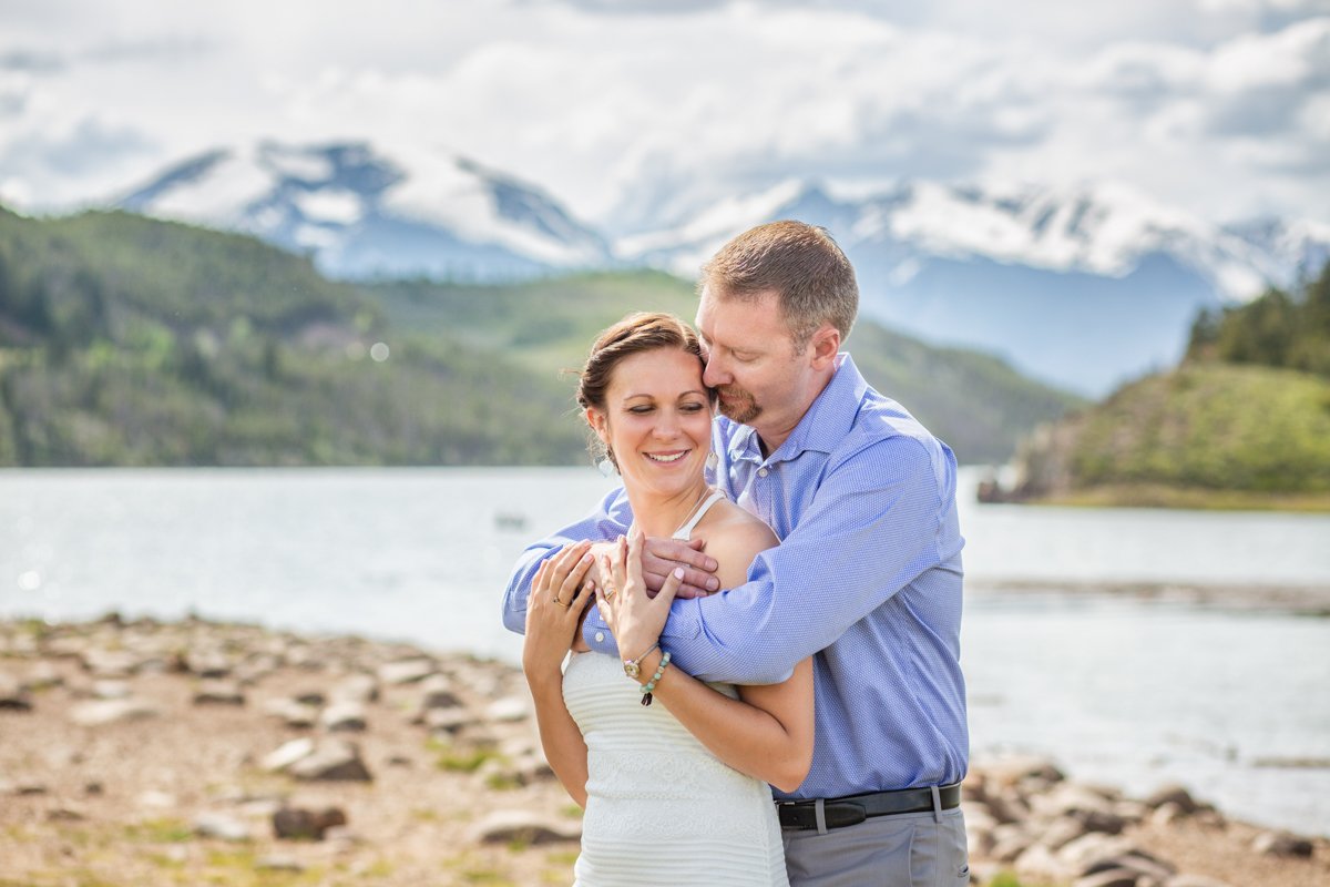 A couple embrace along the shoreline of a lake with snow capped mountains in the back.