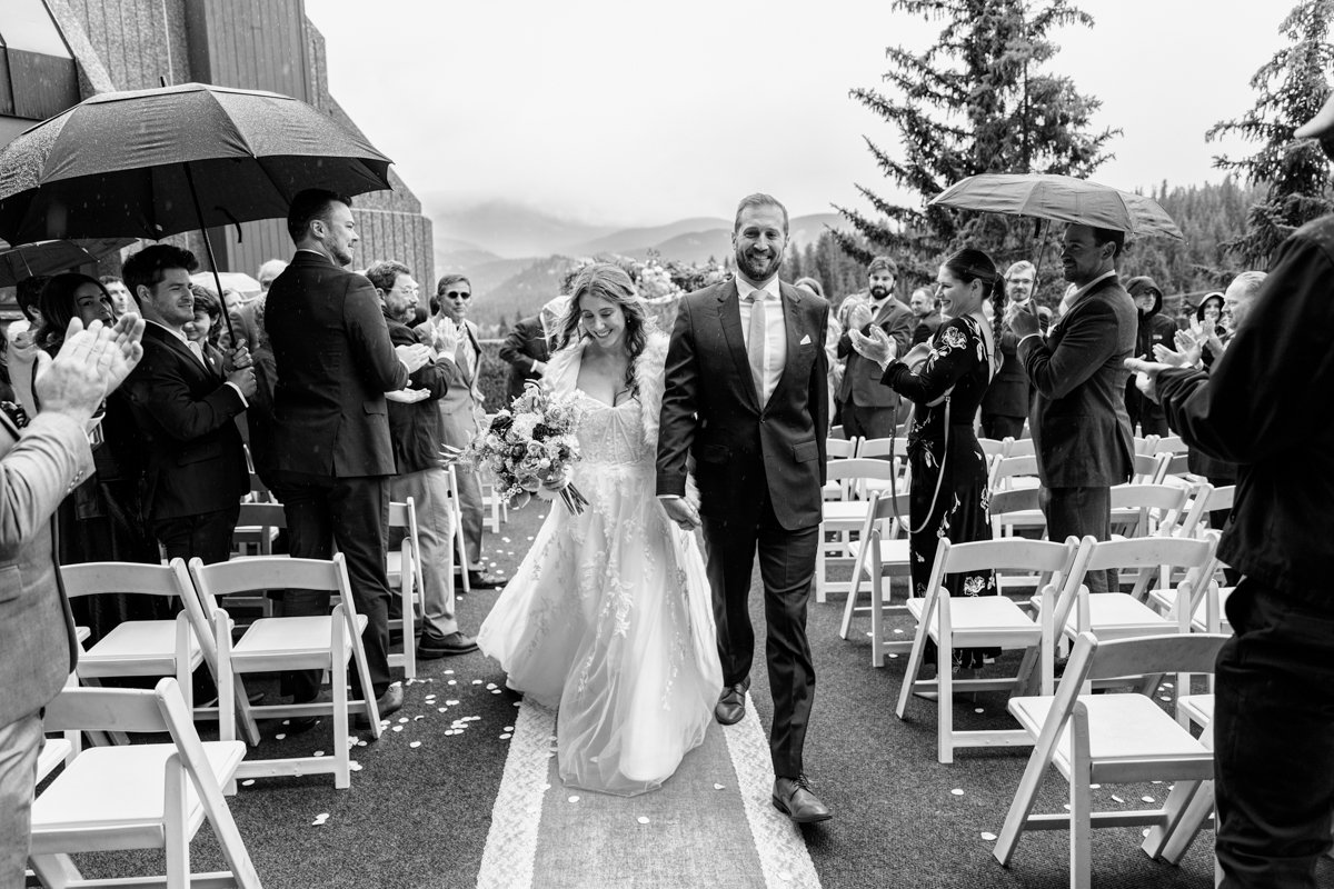 A couple smiles as they walk down the aisle after their vows. Black and white image. It's raining and the guests have umbrellas. Misty mountains in the background with storm clouds.