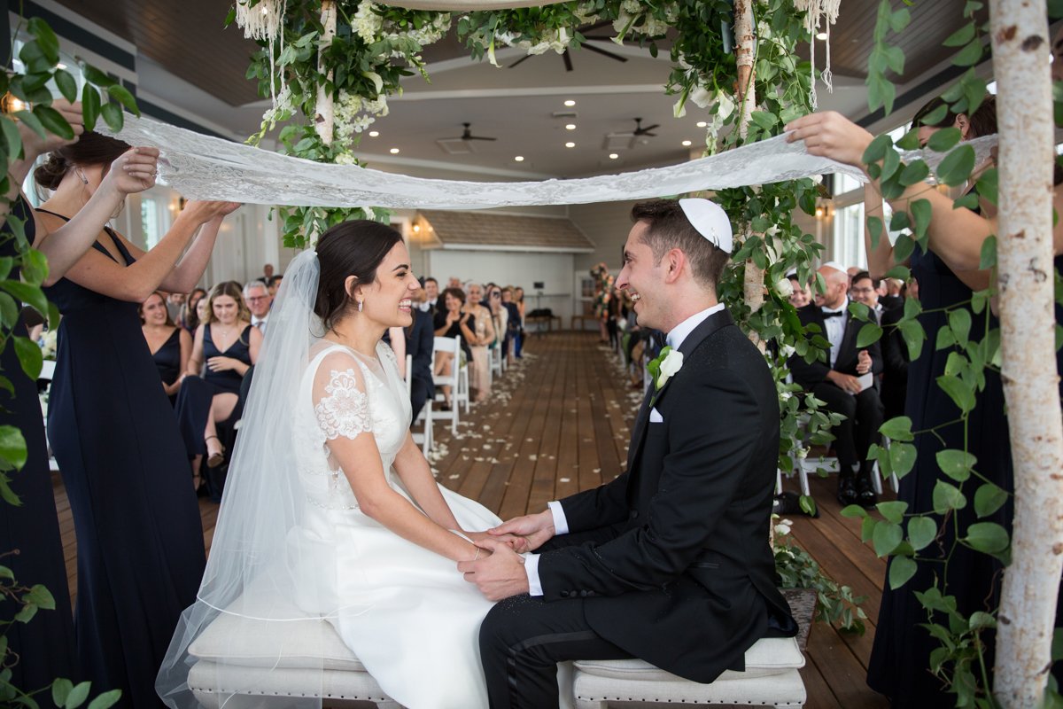 A couple sit during their fusion ceremony. They hold hands under a Persian Sugar Cloth while they sit under a chuppah.