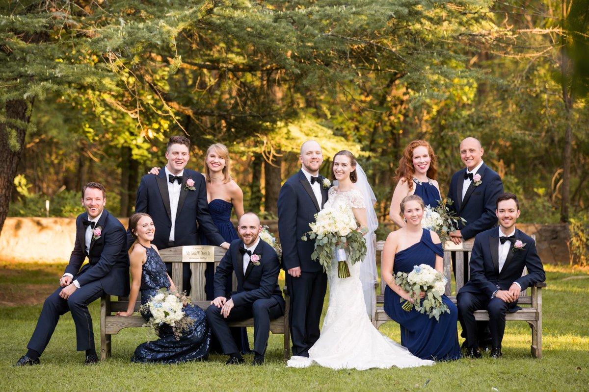 Formal and fun bridal party photo. The couple stands in the center. The bridal party is situated on either side, some sitting on benches, some standing.