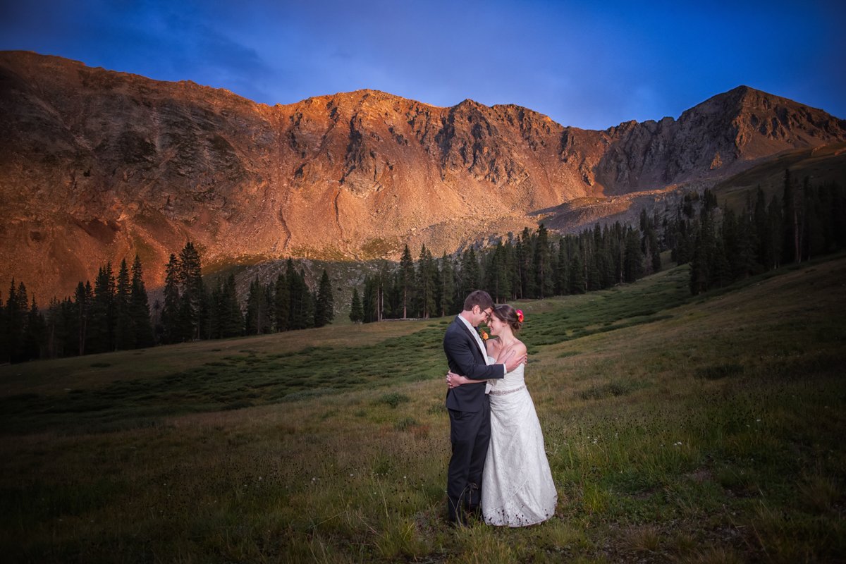 Couple embraces at sunset on top of Arapahoe Basin. Large mountain rock wall is illuminated with alpenglow.