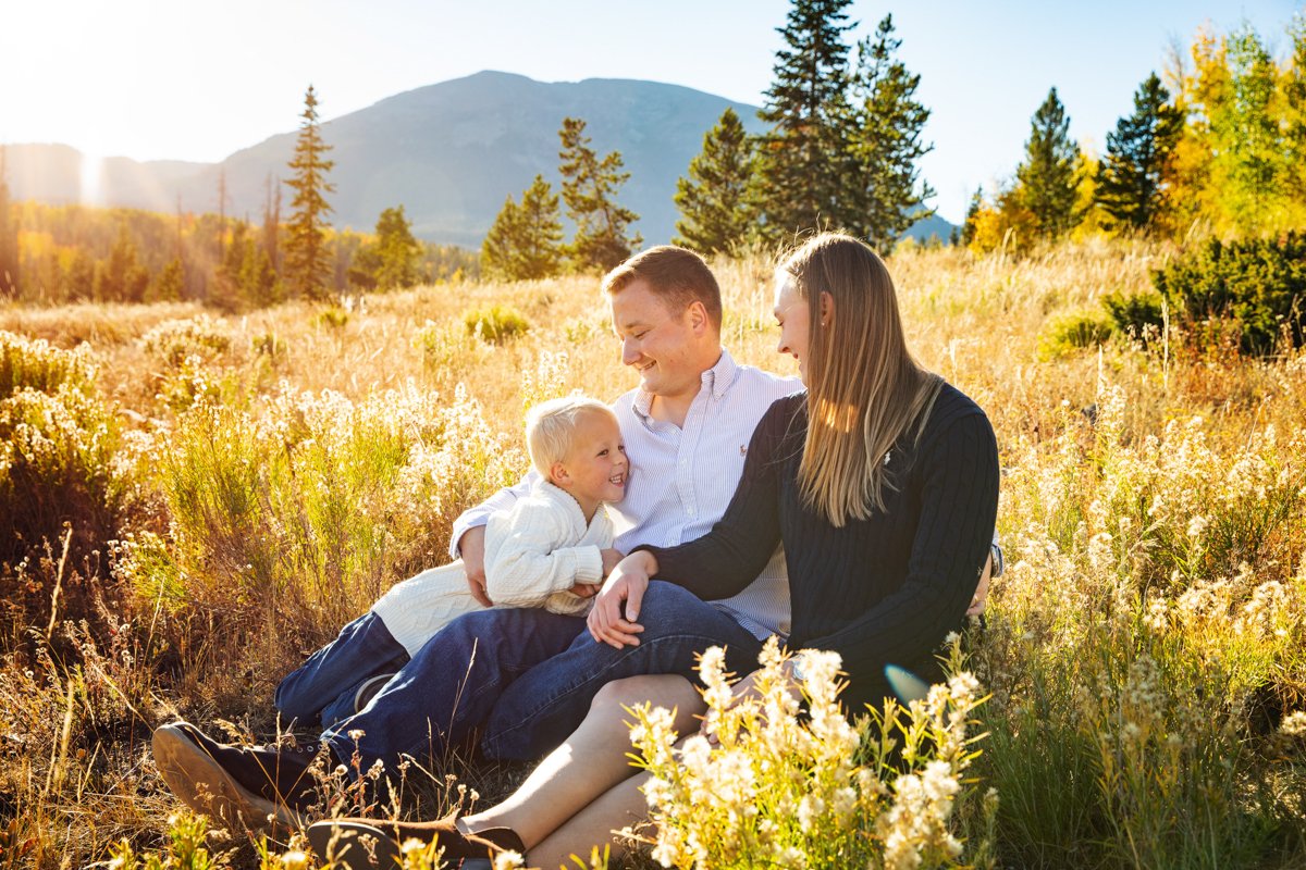 Young couple sits in a field at sunset with their young son on their laps. Mountains in the background