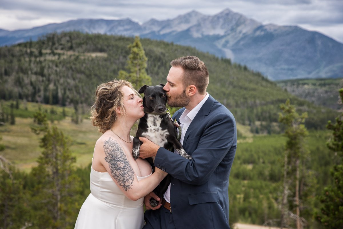 A tattooed bride and her groom with a mountain backdrop pick up and hold their dog and they both kiss the dog on the cheeks at the same time.