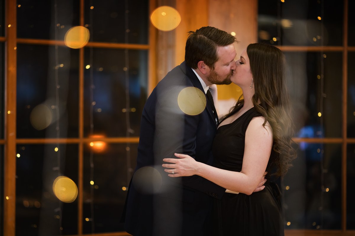 Non traditional couple wears black wedding dress. They kiss as they're layered between twinkling lights in the camera frame.