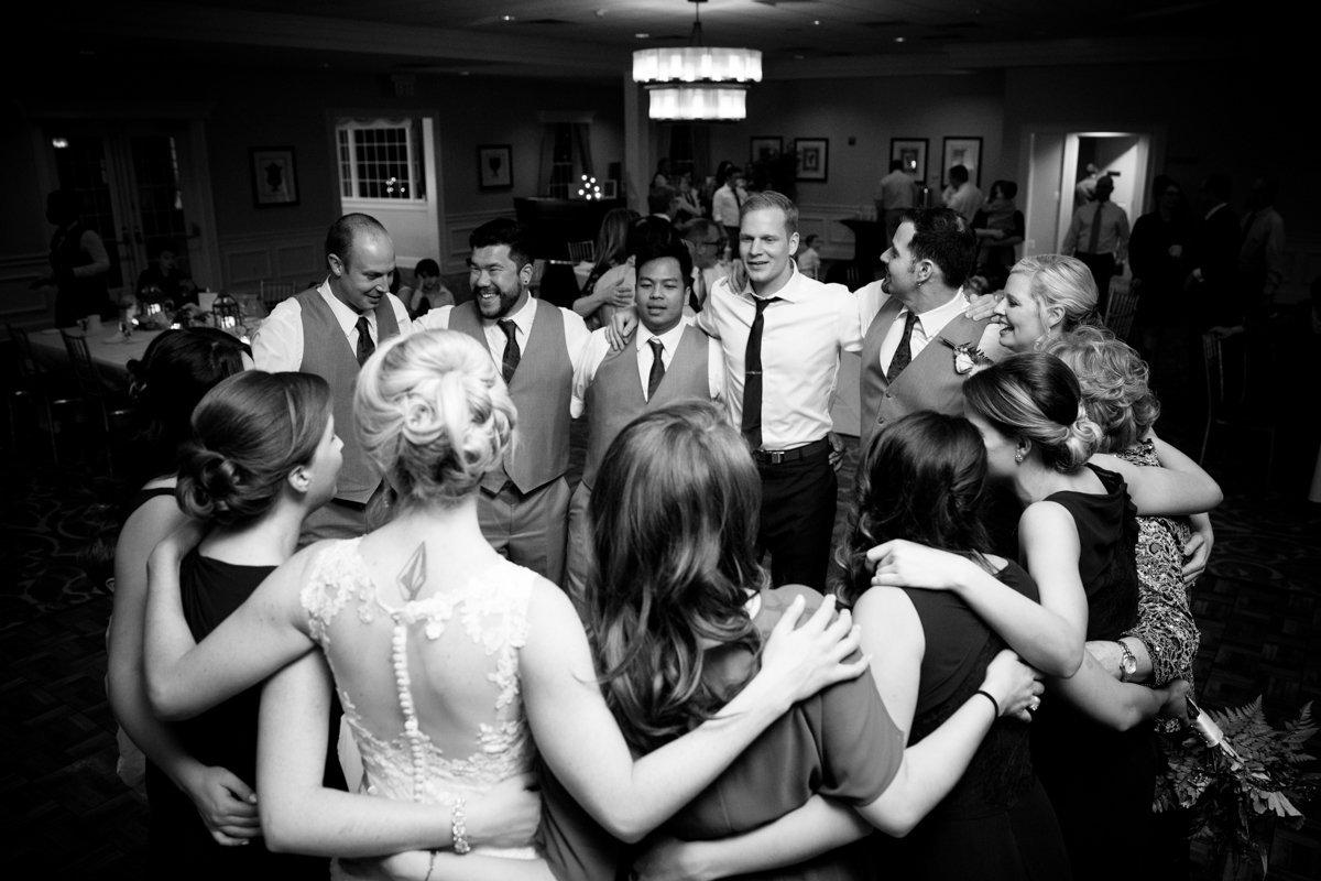 Bride and groom on the dance floor with arms around all of their friends in a circle.
