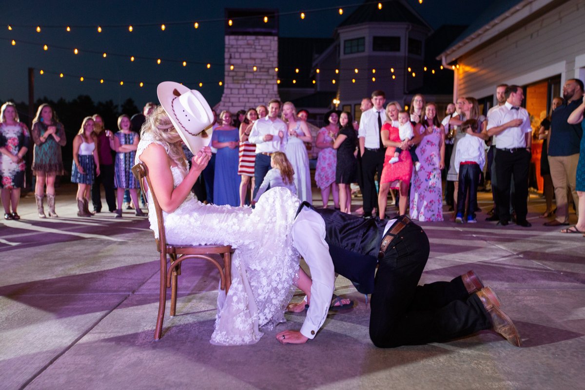 Bride sits in a chair on the dance floor. she wears a cowboy hat and covers her face as her groom goes under her dress for the garter in the traditional garter toss.