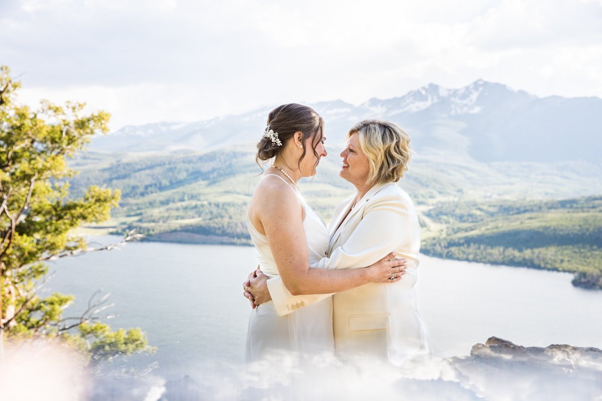 Two brides embrace on the top of a mountain with peaks behind them. The sky is artistically reflected in a prism.