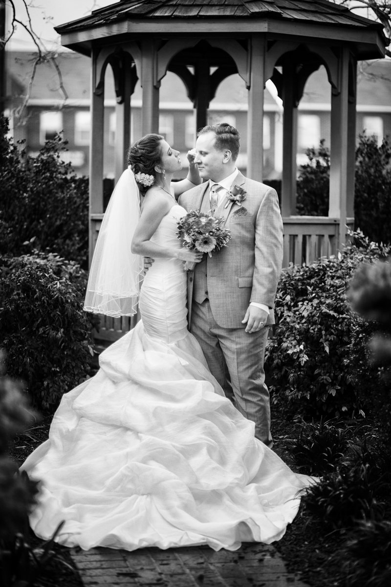 Casual moment between photos where the bride her her dramatic fit n flair dress fixes the hair of her groom as they stand in a garden in front of a gazebo..