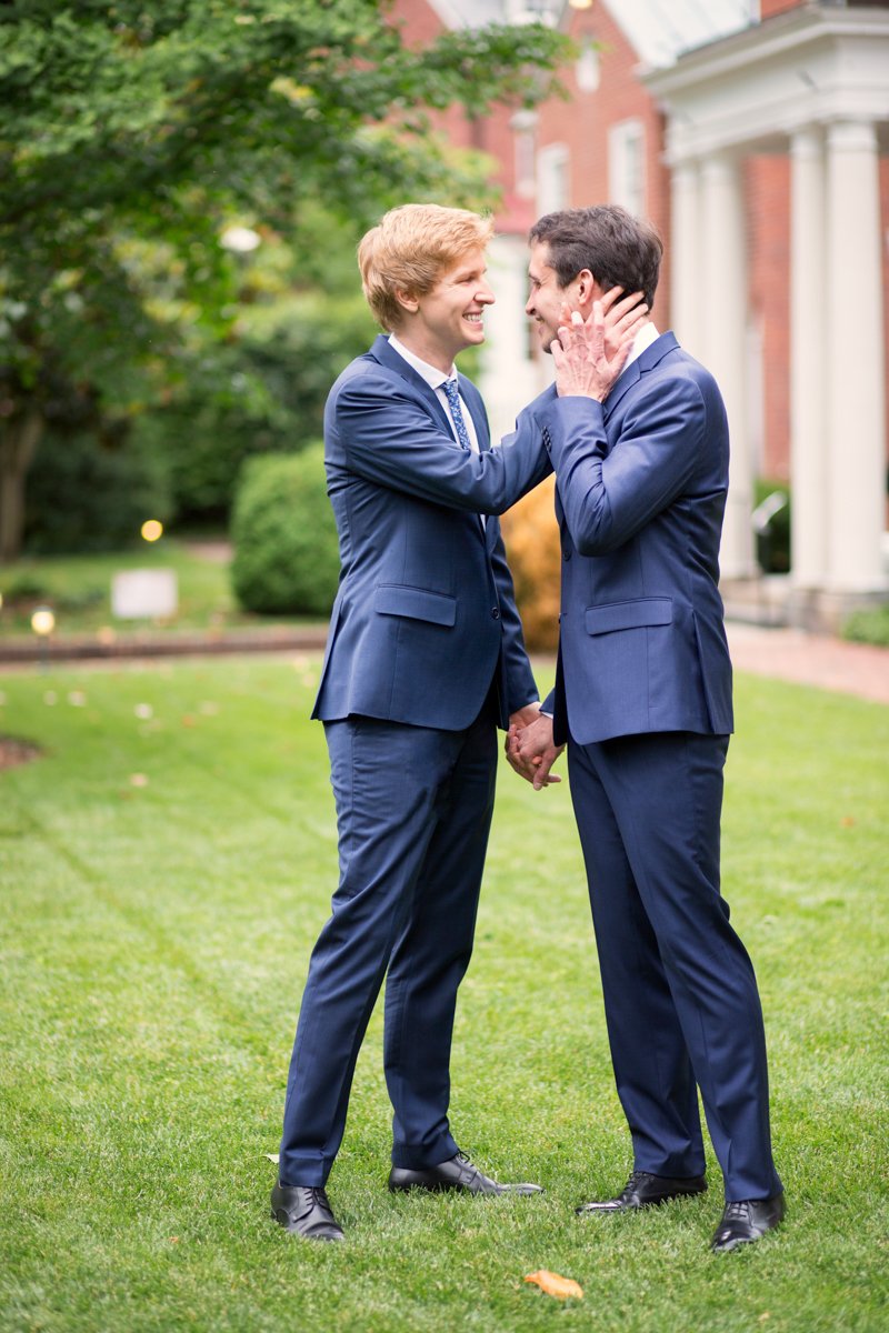 Two grooms share a moment on the lawn of their wedding venue. They hold hands and one groom touches the other's neck.