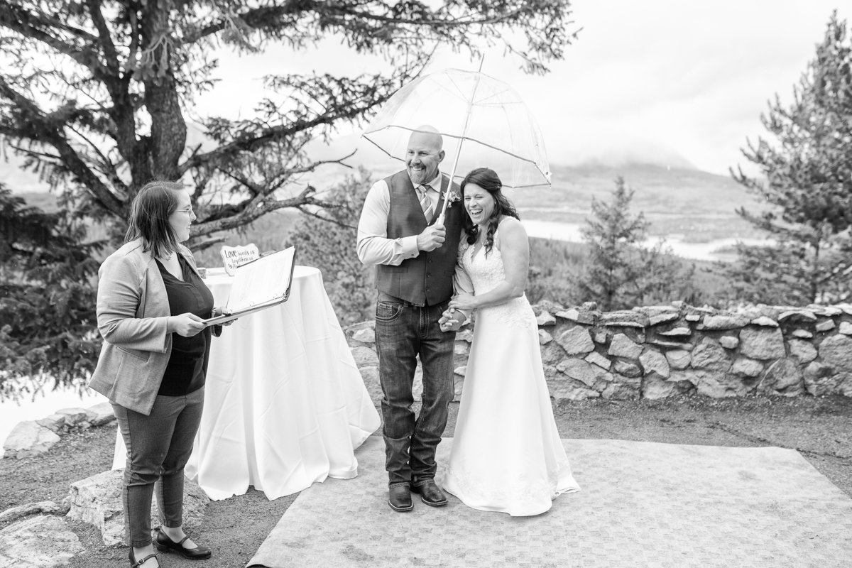 Black and white photo of a couple laughing at their elopement ceremony while they're under a clear umbrella as it snows and rains in the summer at Sapphire Point.