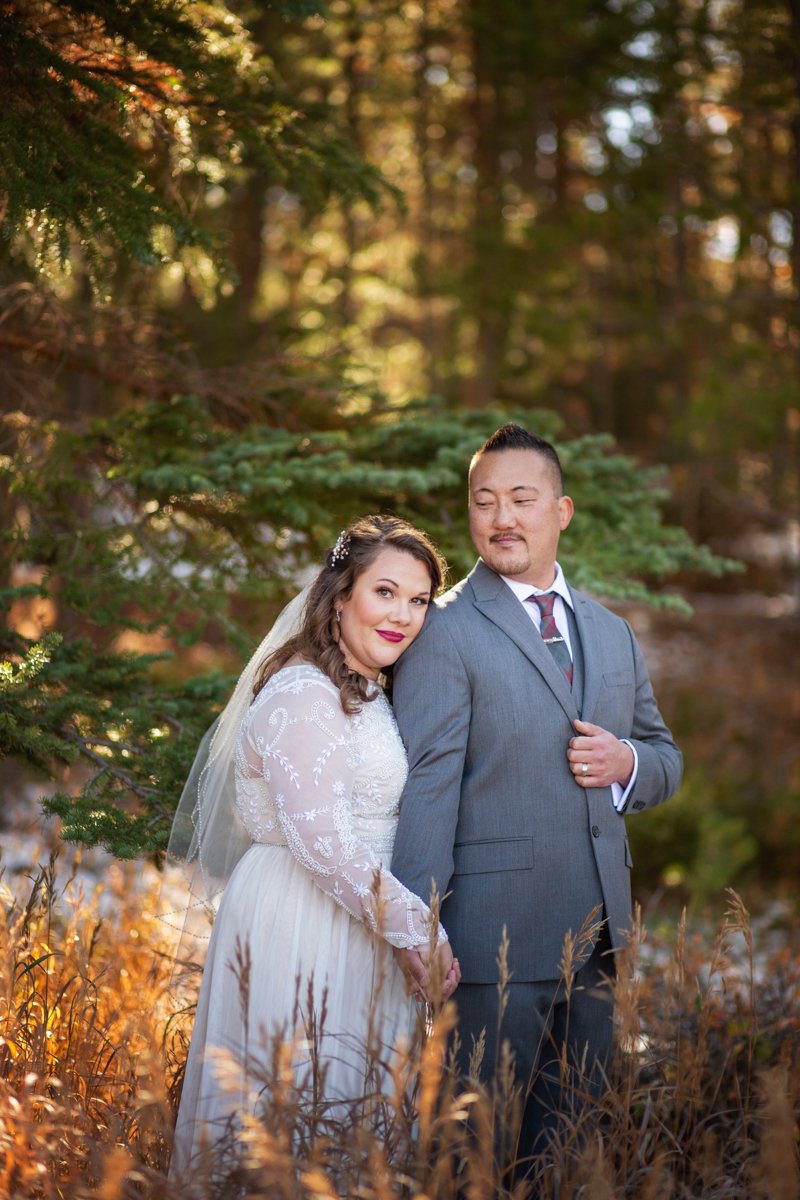 A groom looks dapper in a grey suit and holds the hand of his bride in a dress with lace sleeves and a veil as she rests her cheek on his shoulder.
