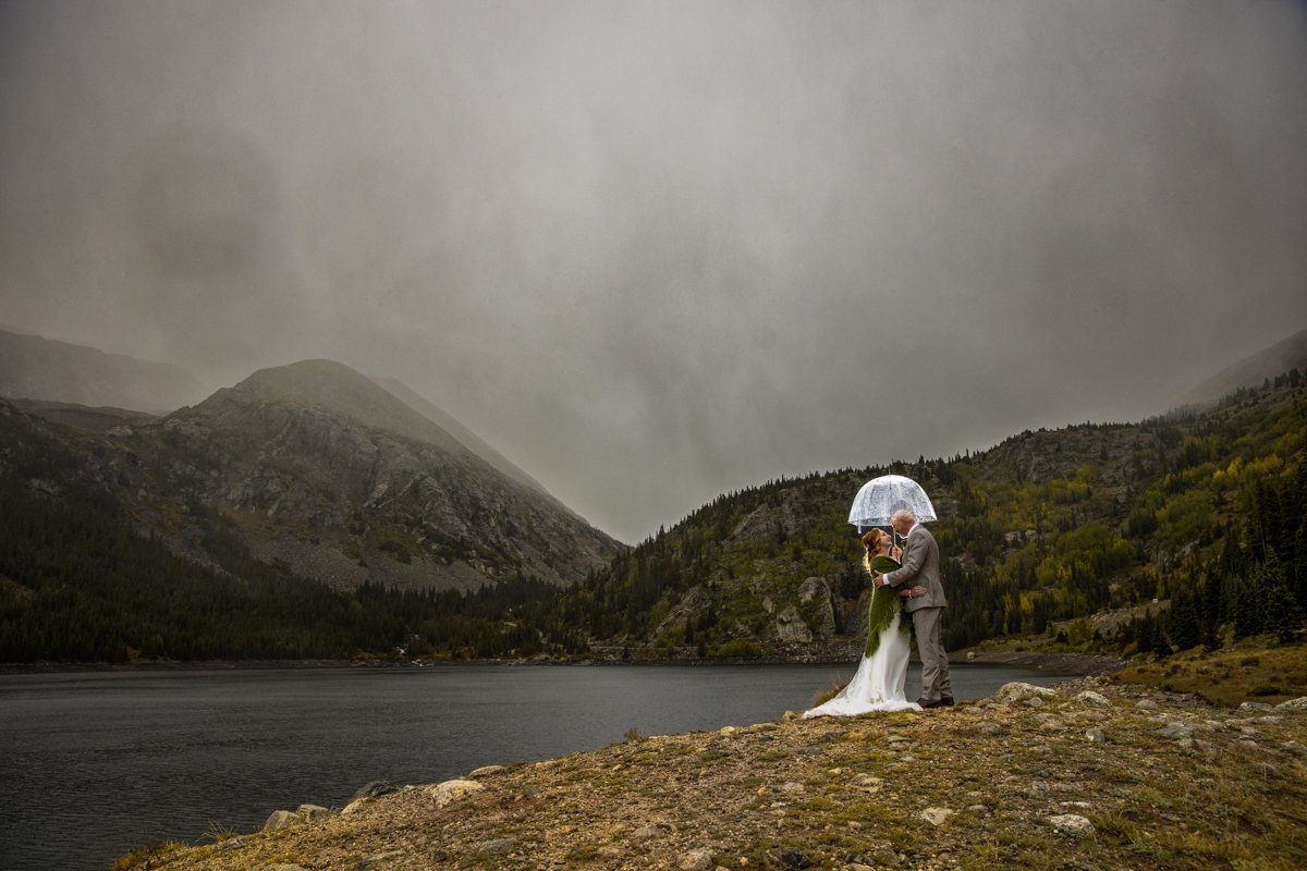 Photo of a couple snuggled together under an umbrella in the rain along the shoreline of a mountain lake.
