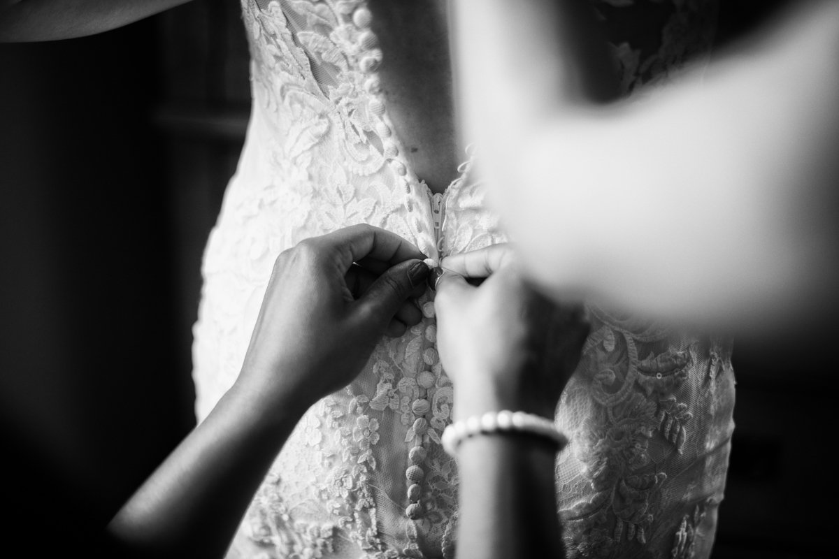 Classic detail photo in black and white of a bride getting her dress buttoned up the back. Just a detailed/close up of hands working the buttons.