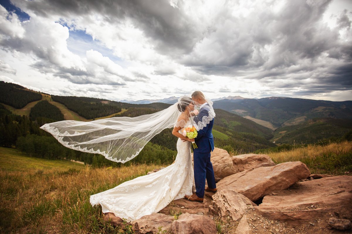 Couple stand on boulders. Bride's veil is over the couple and also trails off behind her in the wind. lush green mountains and dramatic clouds in the background.