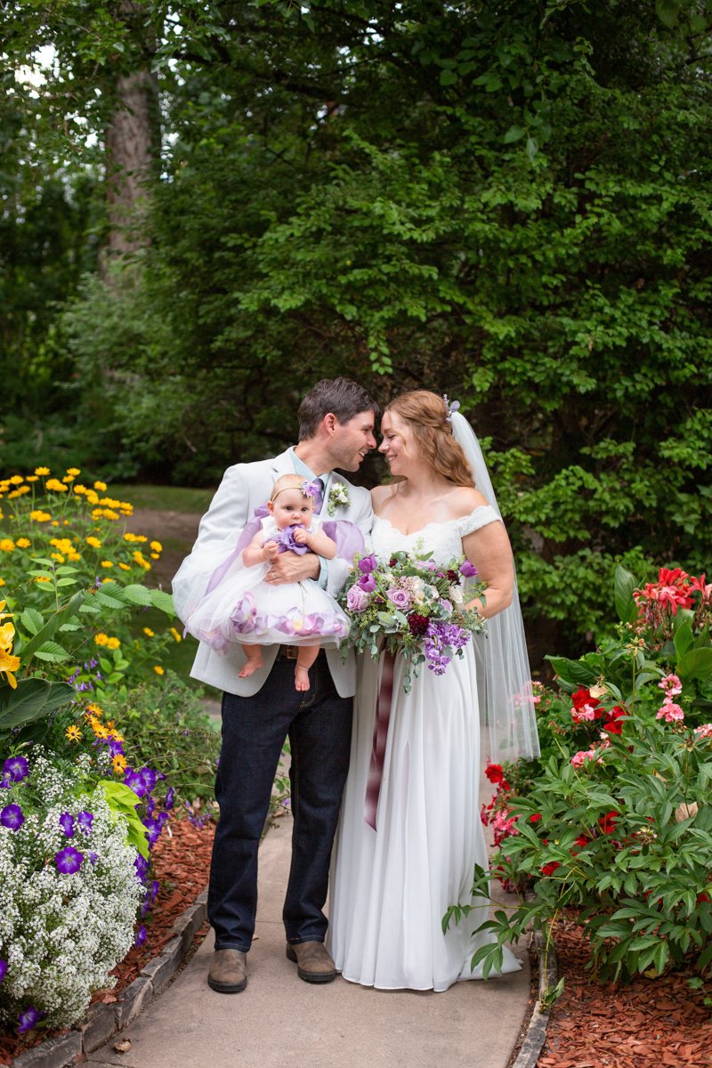 A couple stand in a garden during their wedding day holding their infant baby daughter.