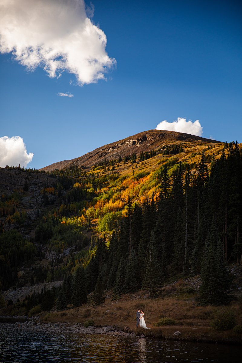 A far away photo of a couple on a lake shore, illuminated in a light beam, with golden trees and evergreen mountains behind them in the Fall.