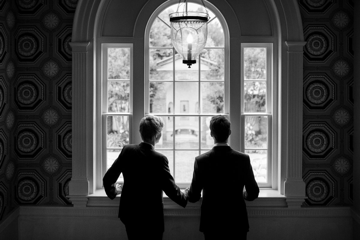 Two grooms in silhouette looking out a window at their ceremony spot.