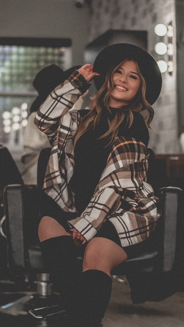 A woman wearing a black hat, plaid jacket, and black thigh-high socks, sitting in a salon chair, smiling and adjusting her hat.