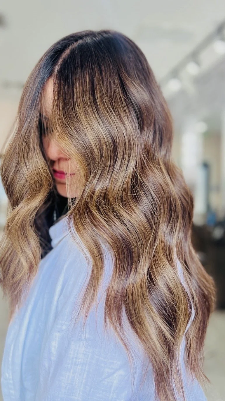 A woman with long, wavy hair styled in loose waves with a balayage coloring effect, featuring darker roots and lighter ends, in a salon setting.