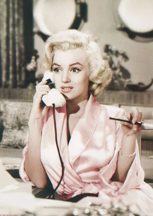 A woman with blonde, vintage-style hair wearing a pink satin robe, speaking on a corded telephone with a concerned expression, holding a makeup brush in her other hand, in a room with decorative plates on the wall.