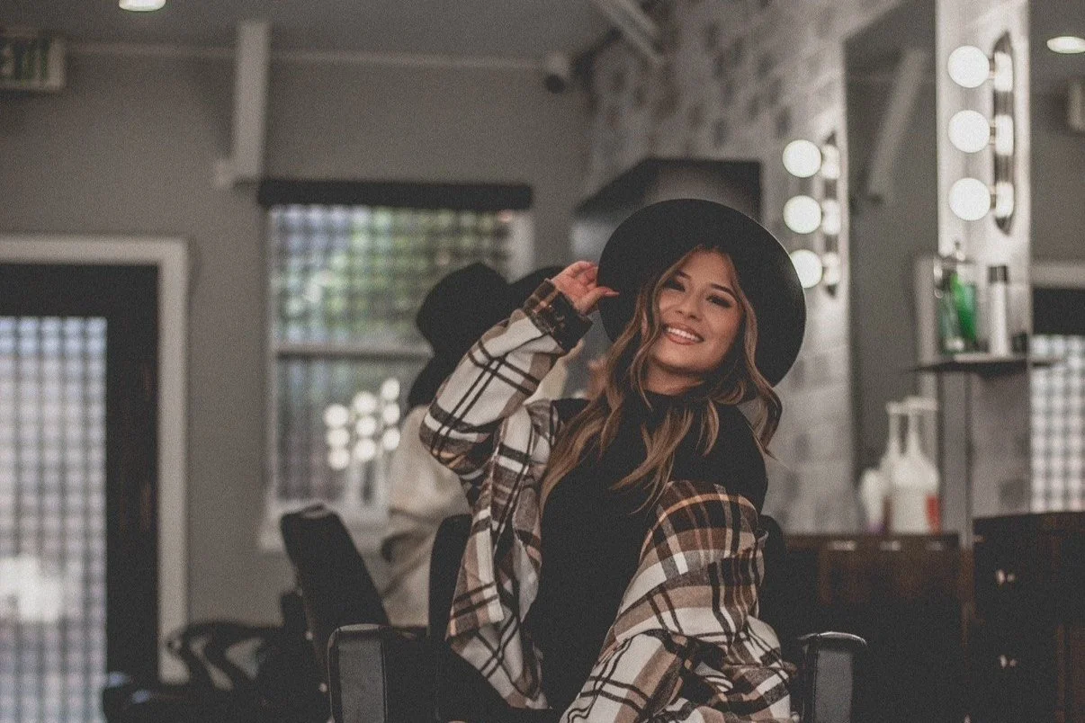 A smiling woman wearing a black hat and a plaid jacket, sitting in a salon or dressing room, with a makeup mirror and lights in the background.