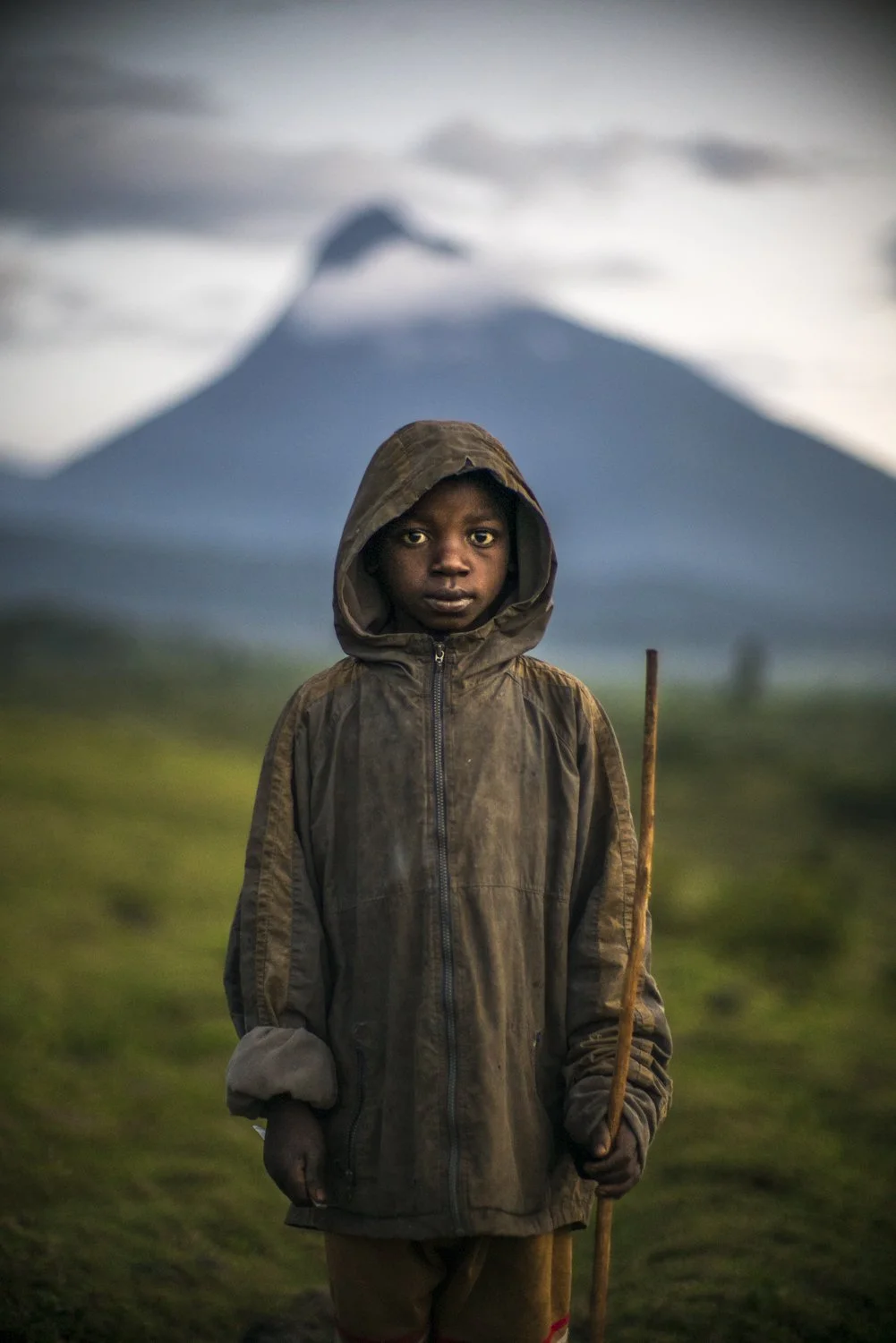 A young boy in a worn, hooded jacket holding a stick, standing outdoors with a blurred volcano in the background.