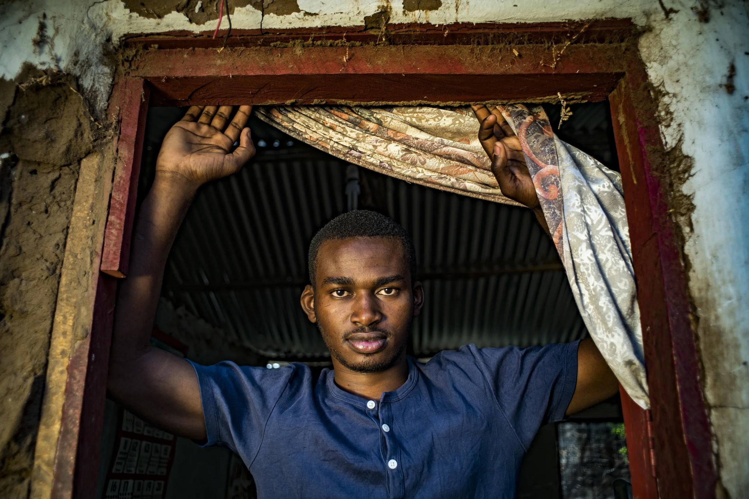 A young man stands inside a doorway framed by a rough wall, holding back a curtain with both hands. He has a serious expression and is wearing a blue shirt.