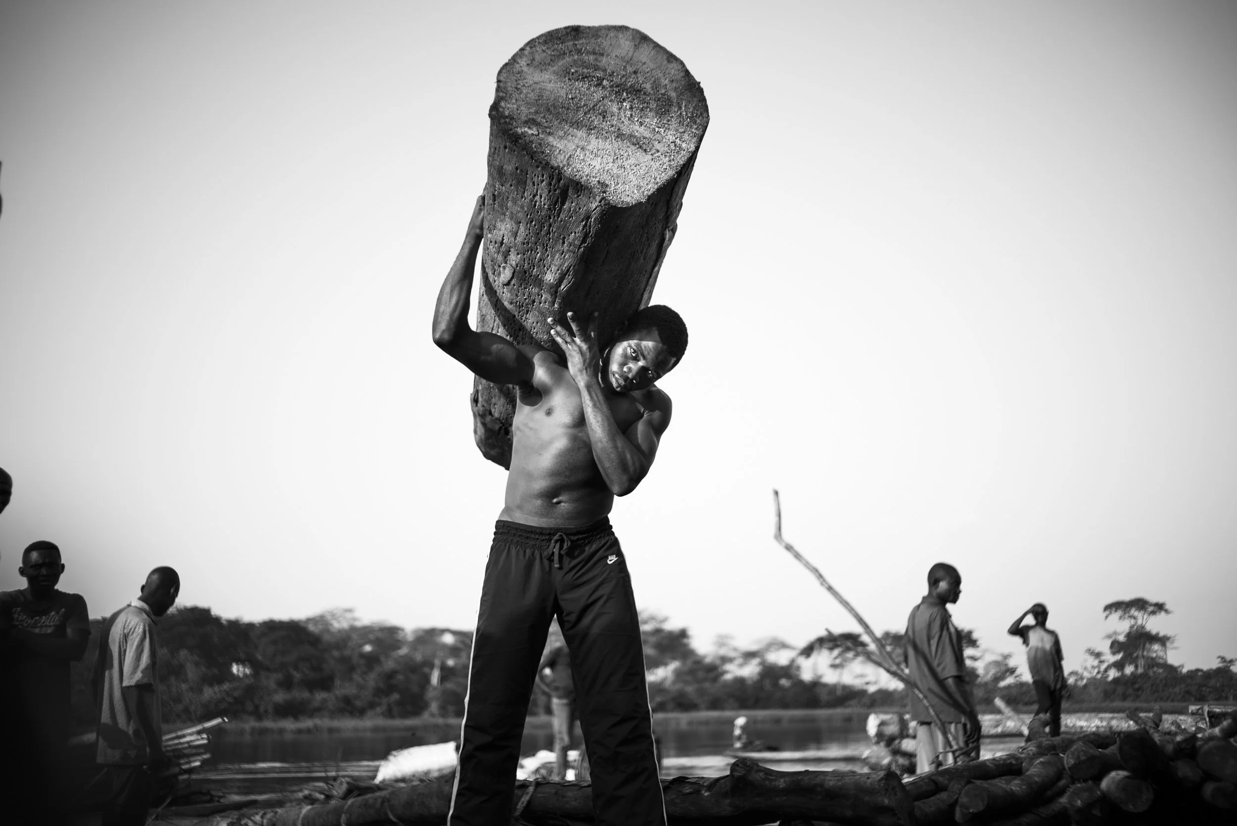 A shirtless man carrying a large log on his shoulder near a river with other men in the background.