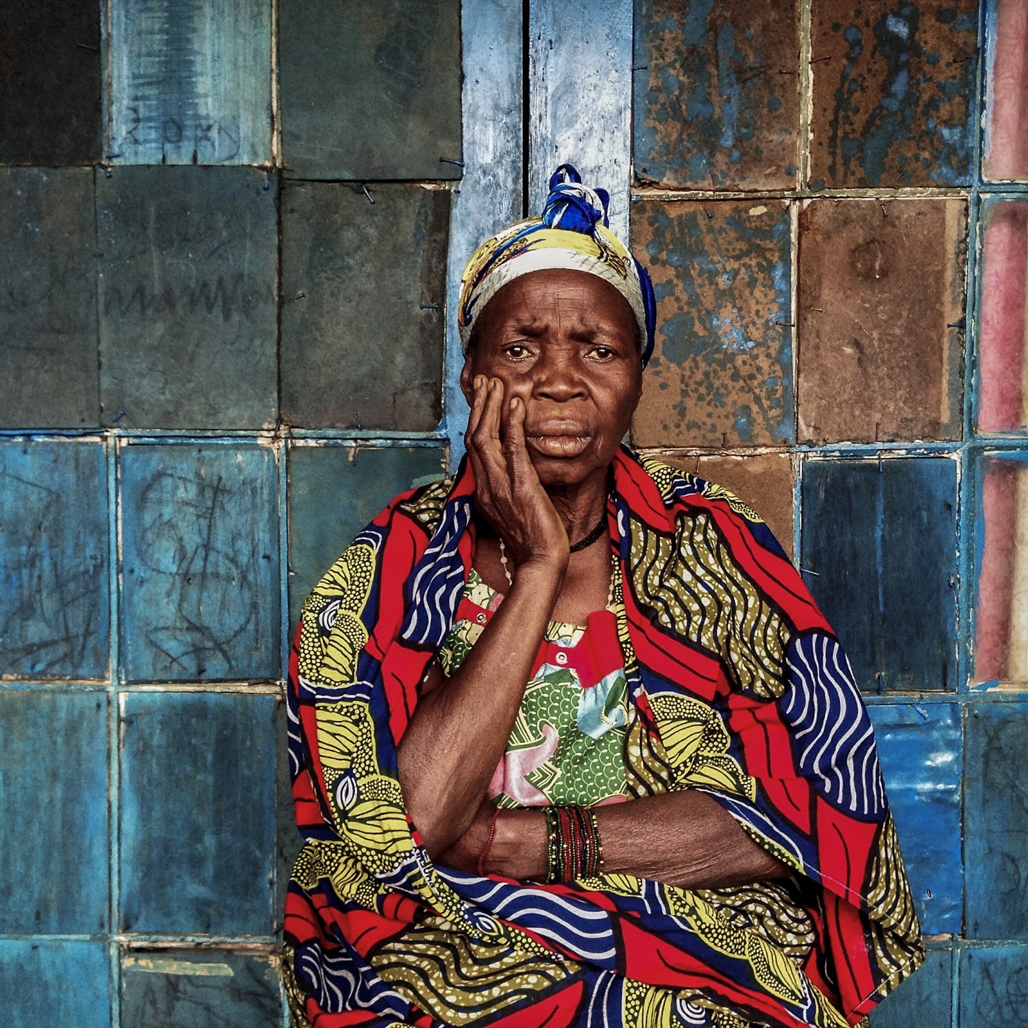 An elderly woman with a concerned expression, resting her face in her hand, standing against a colorful tiled wall.