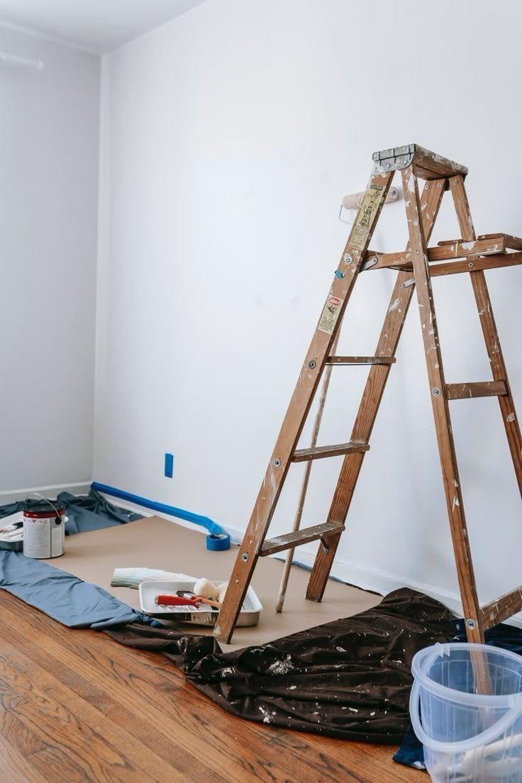 Room interior with a wooden ladder, paint supplies, and drop cloths on a hardwood floor, prepared for painting.