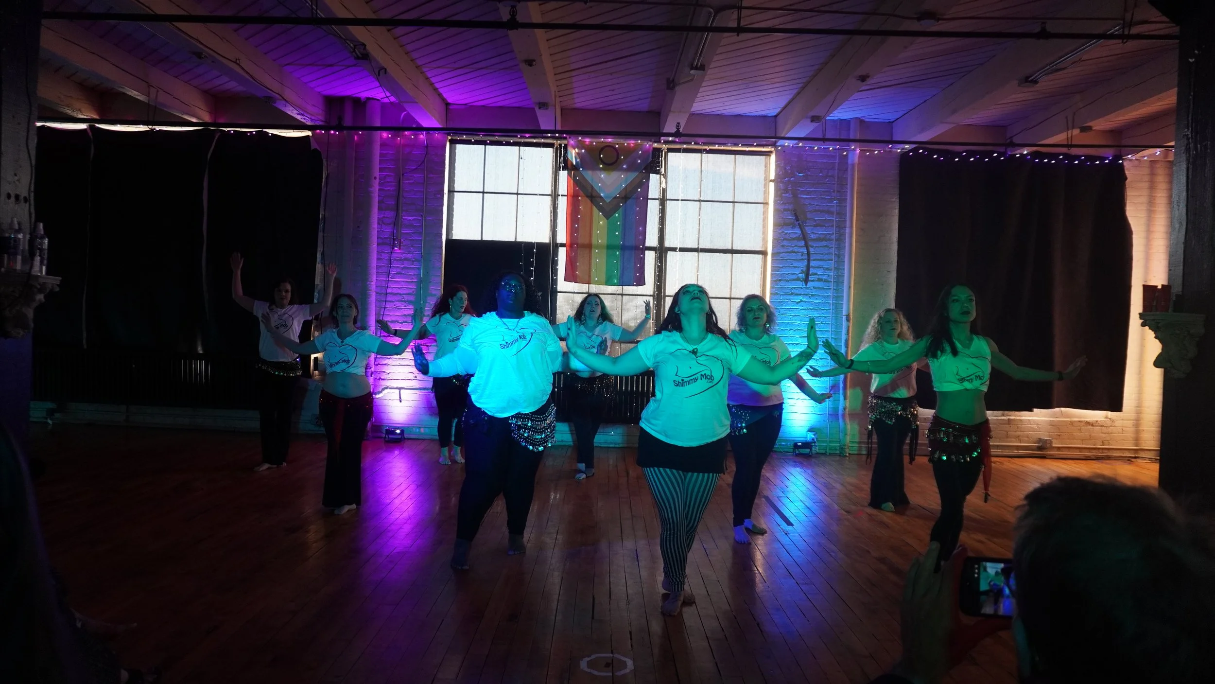 Group of women performing a dance routine on stage, wearing matching white T-shirts with a logo, in an indoor venue decorated with purple and blue lighting and an LGBTQ+ rainbow flag hanging in the background.
