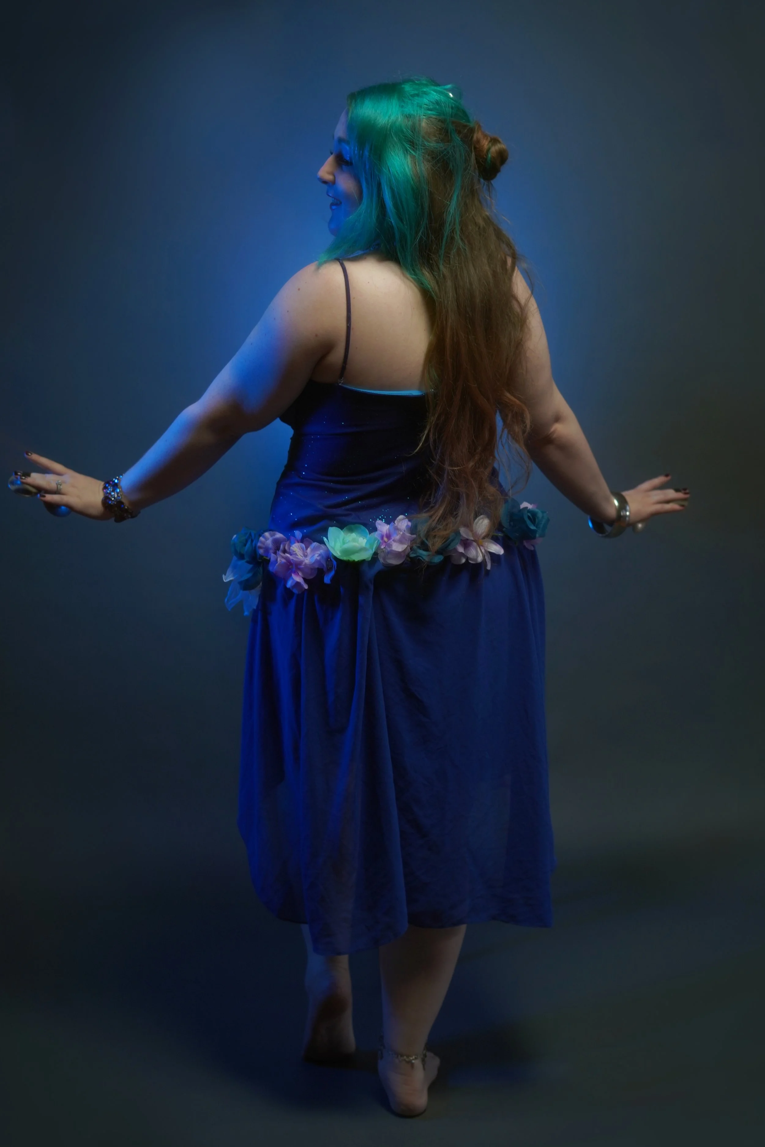 A woman with long, wavy hair dyed green on top and brown underneath, posing in a dark studio with blue lighting. She is wearing a dark dress with a floral arrangement at the waist and is standing on her toes. She has jewelry on her wrists and ankles and is smiling.