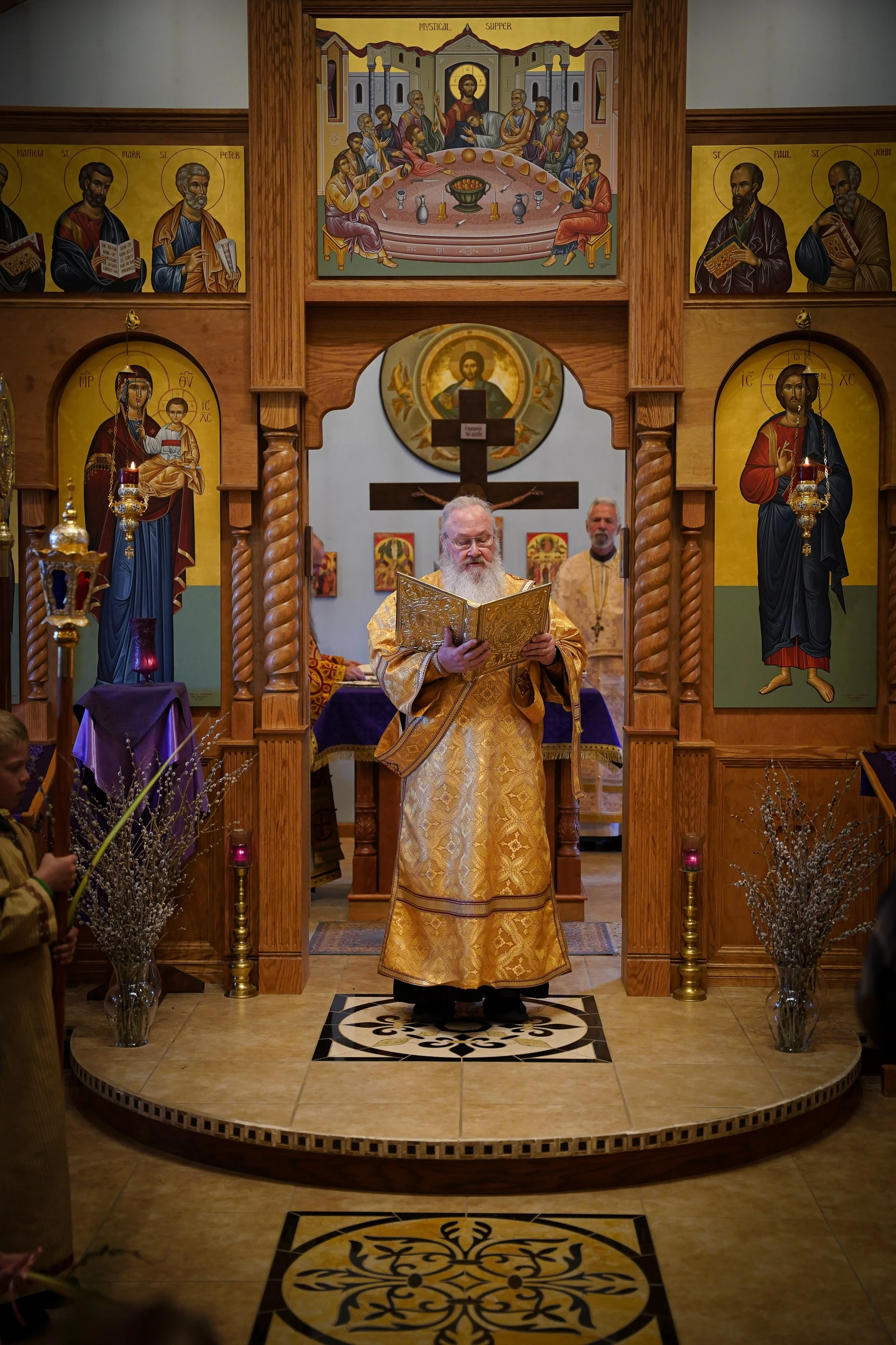 Easter church service with priest in gold vestments reading from a book, standing on a decorated platform. Behind, two men and church icons are visible, including a large icon of Jesus Christ and a large cross.