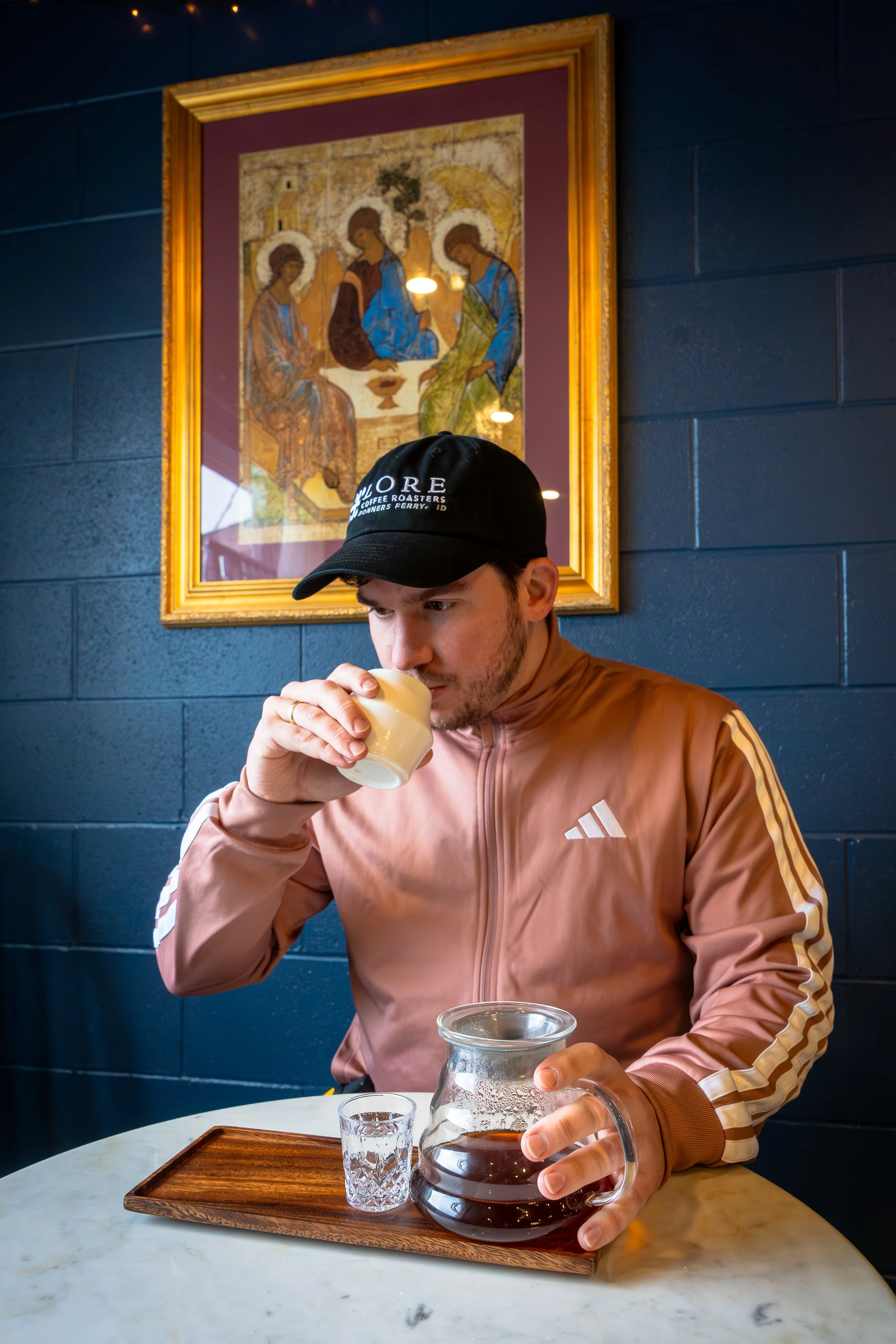 A man wearing a brown Adidas jacket and a black cap sitting at a marble table in a cafe. He is holding a glass of coffee in one hand and pouring coffee from a glass carafe with the other. There is a wooden tray and a glass of water on the table. A fr