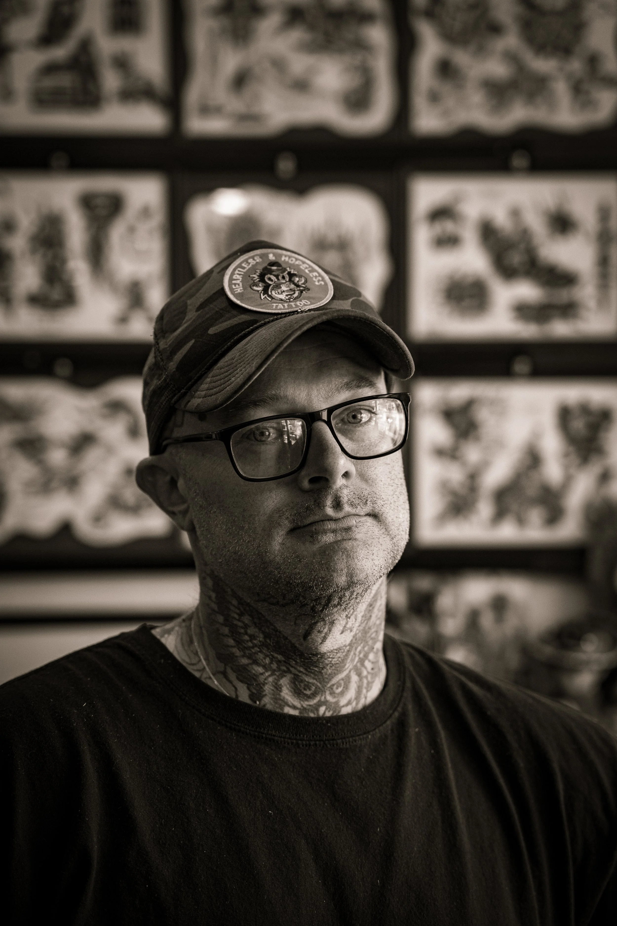 A man wearing glasses, a baseball cap with a patch, and a black shirt, with tattoos on his neck and face, stands in front of a wall decorated with framed tattoo designs.