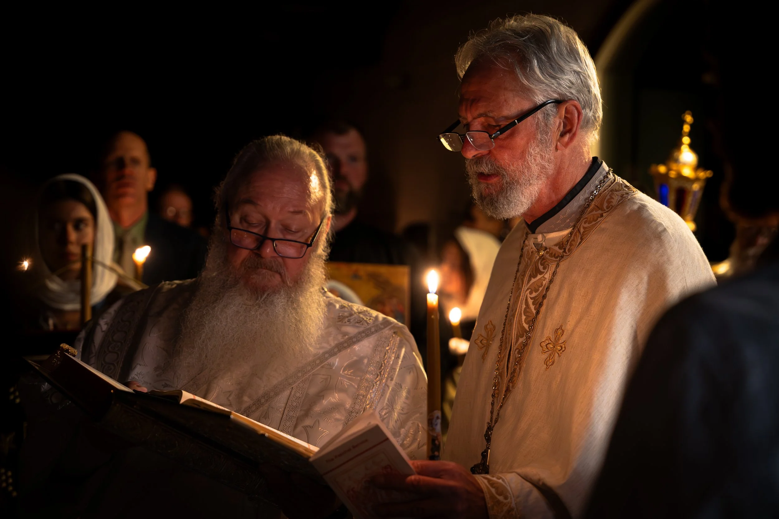 Two elderly men dressed in traditional religious robes reading from a book during a candlelit ceremony, surrounded by other participants.