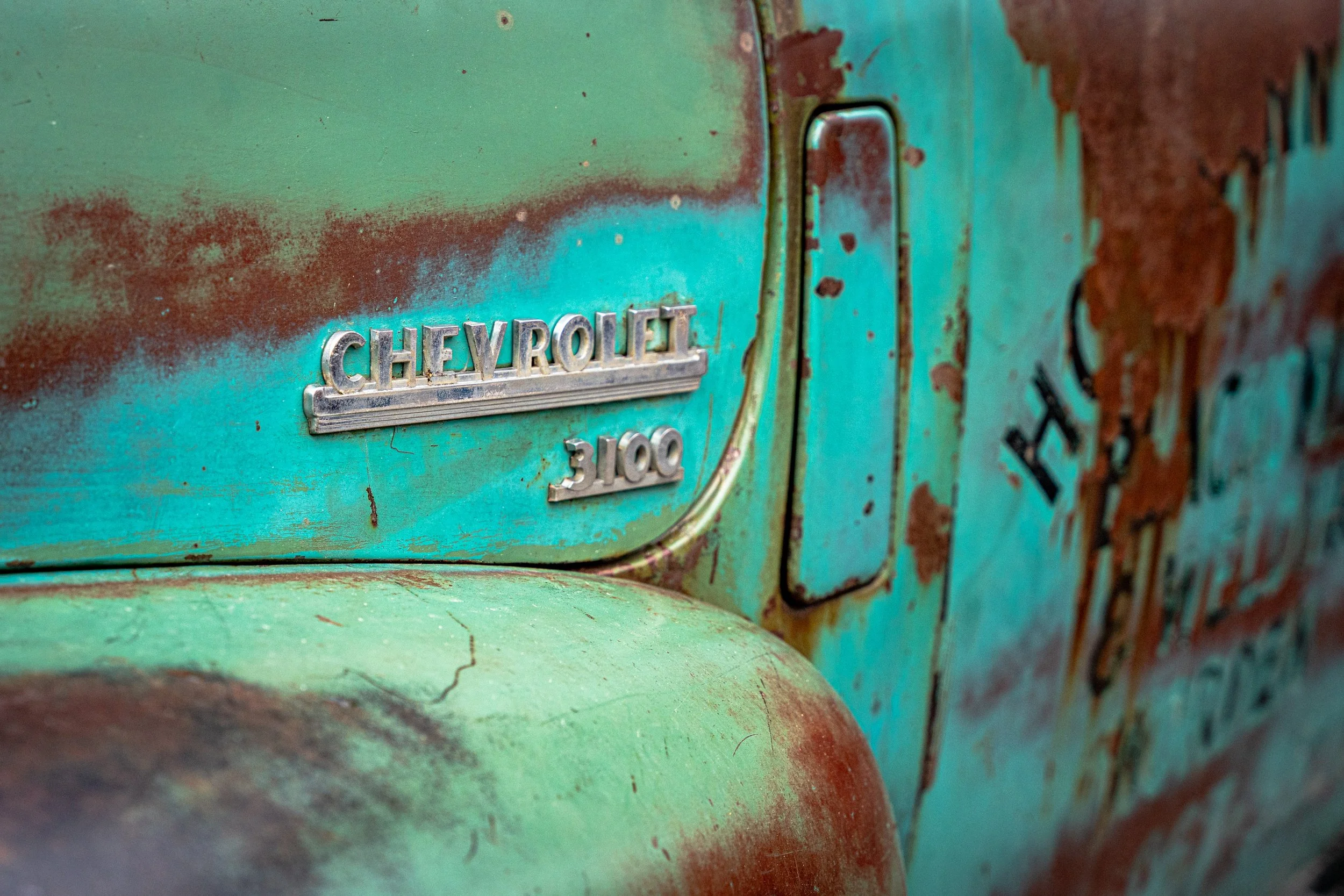 Close-up of an old Chevrolet vehicle with a rusty, weathered teal exterior and a vintage Chevrolet emblem.