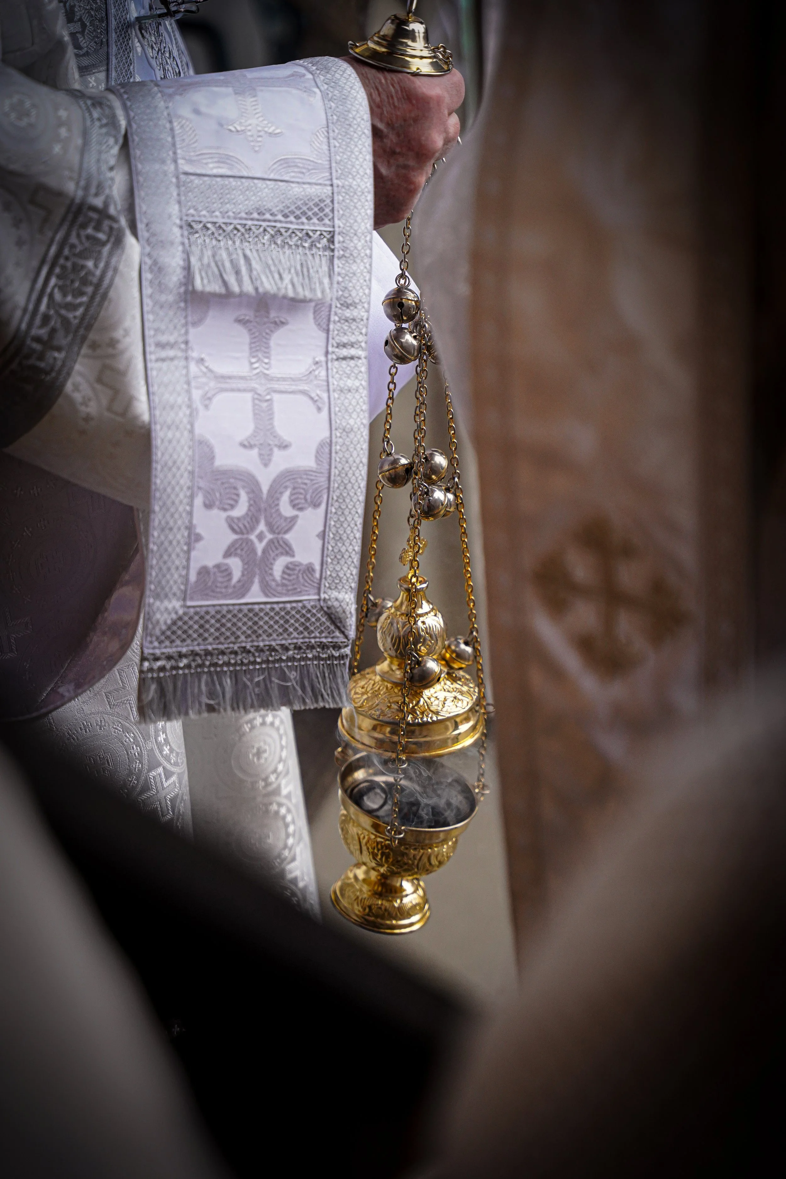 Close-up of a religious ceremonial hand holding a golden thurible, a censers used for burning incense, during a church service, with intricate white vestments and a blurred background.