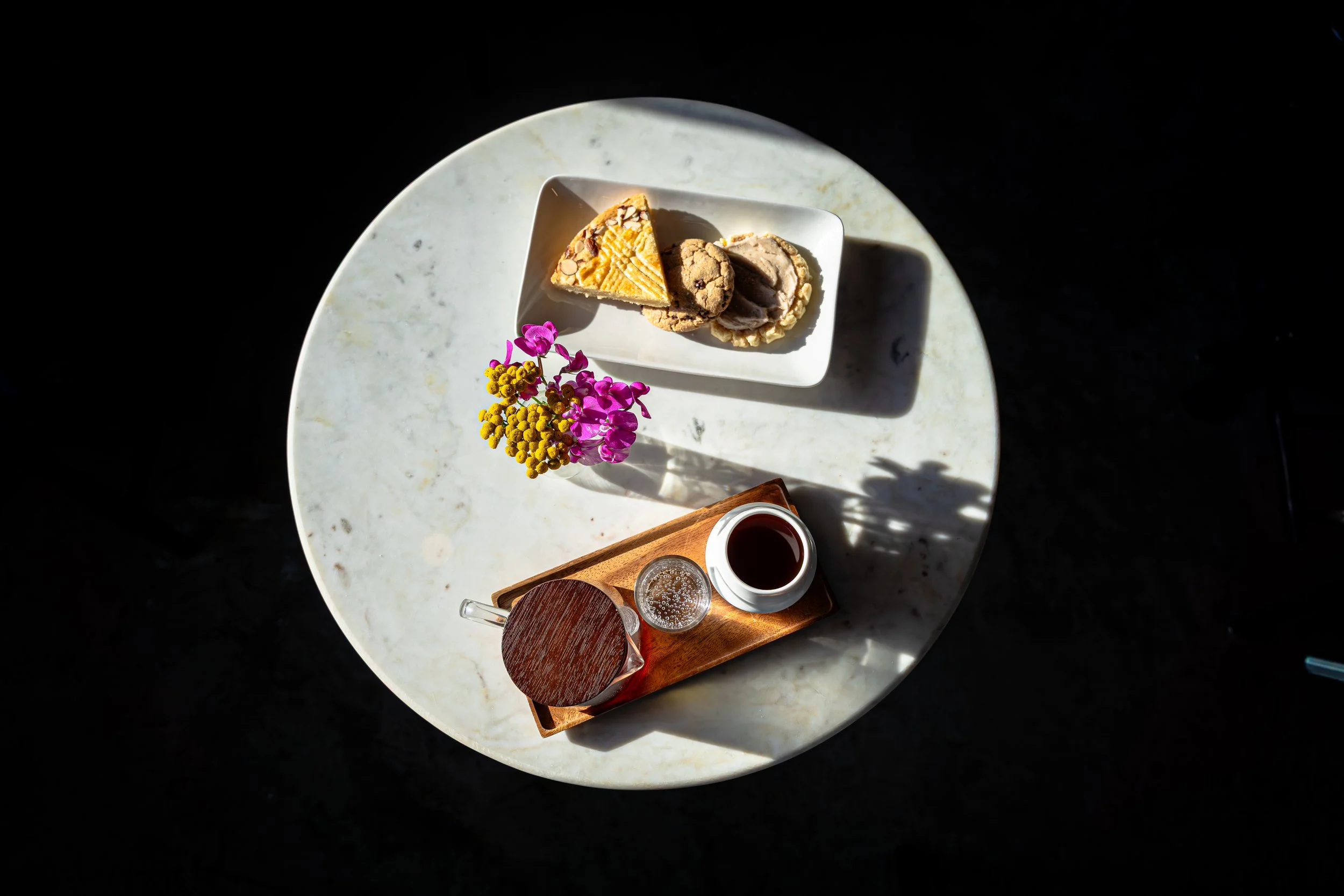 Top-down view of a marble table holding a white rectangular plate with cookies and a triangle-shaped pastry, a small bunch of pink and yellow flowers, a wooden tray with a glass of water, a small white bowl with a dark liquid, and a glass of clear li