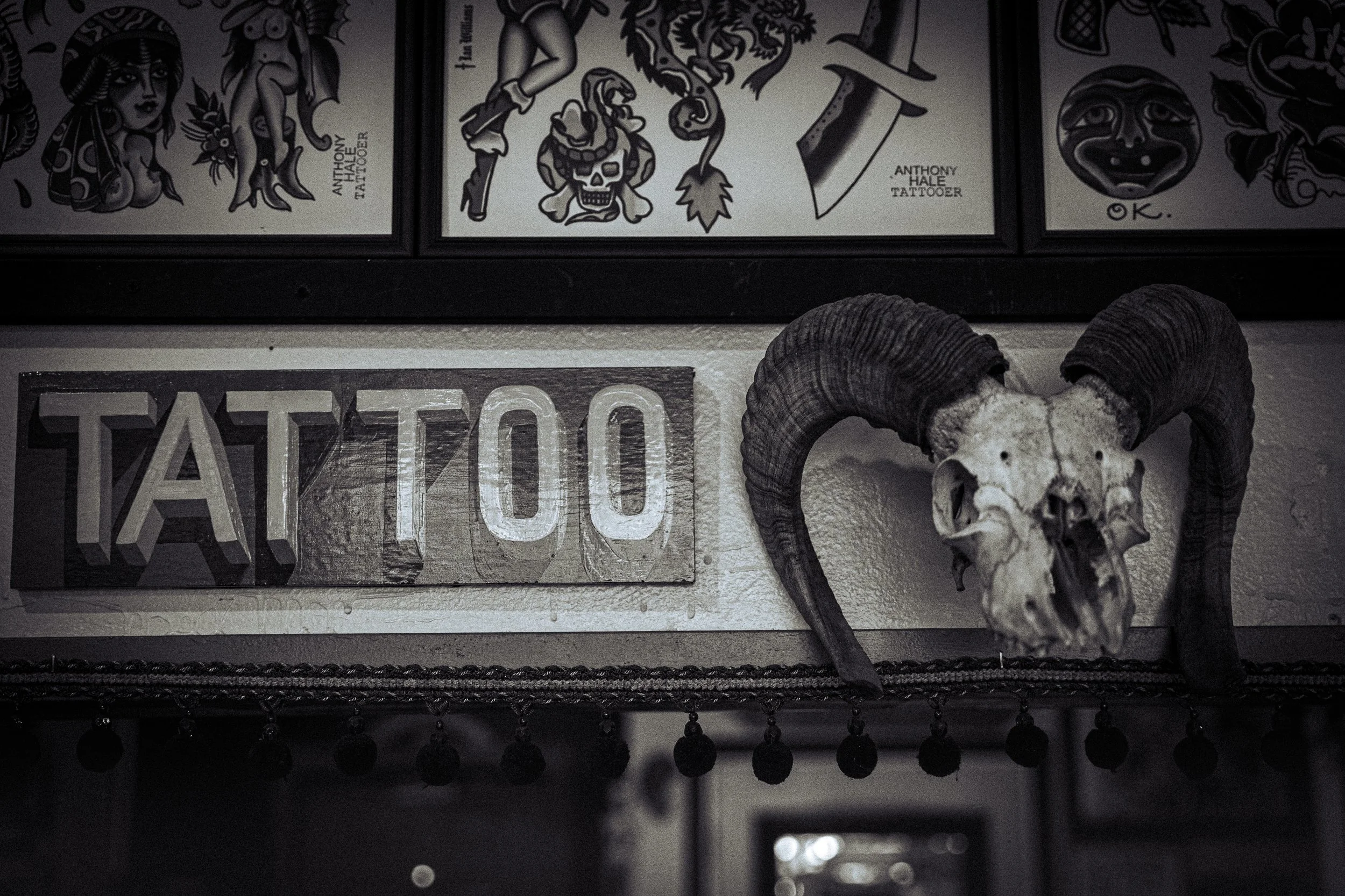 Close-up of tattoo designs on display, including a woman with traditional tattoo art, a skull with a snake, and a face with a smile, accompanied by a ram skull with curved horns in the foreground.