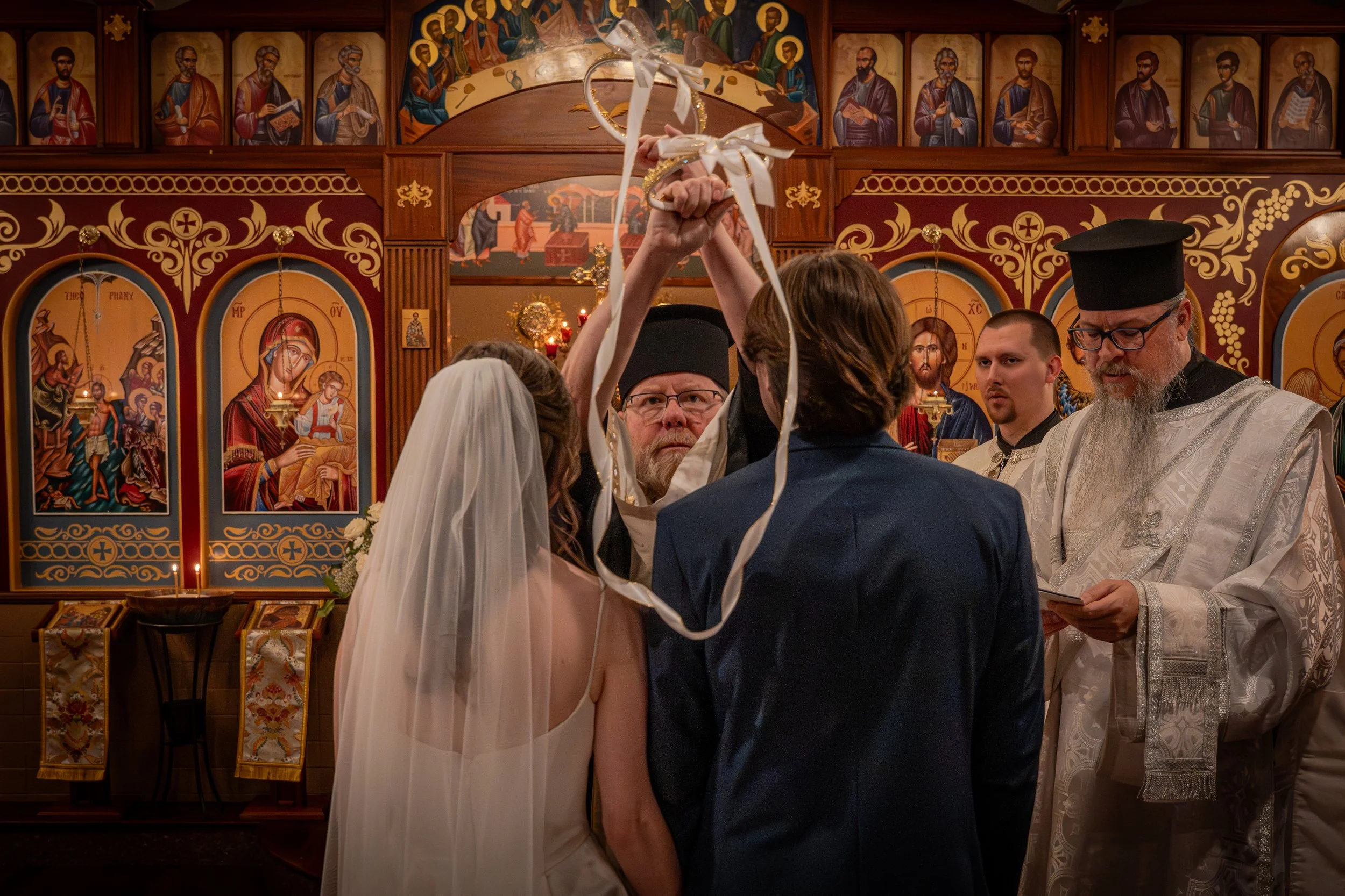 A wedding ceremony inside an Orthodox church with religious icons on the walls, showing a priest in white robes and a black hat, and a bride and groom holding a wedding wreath above their heads, surrounded by clergy and family members.