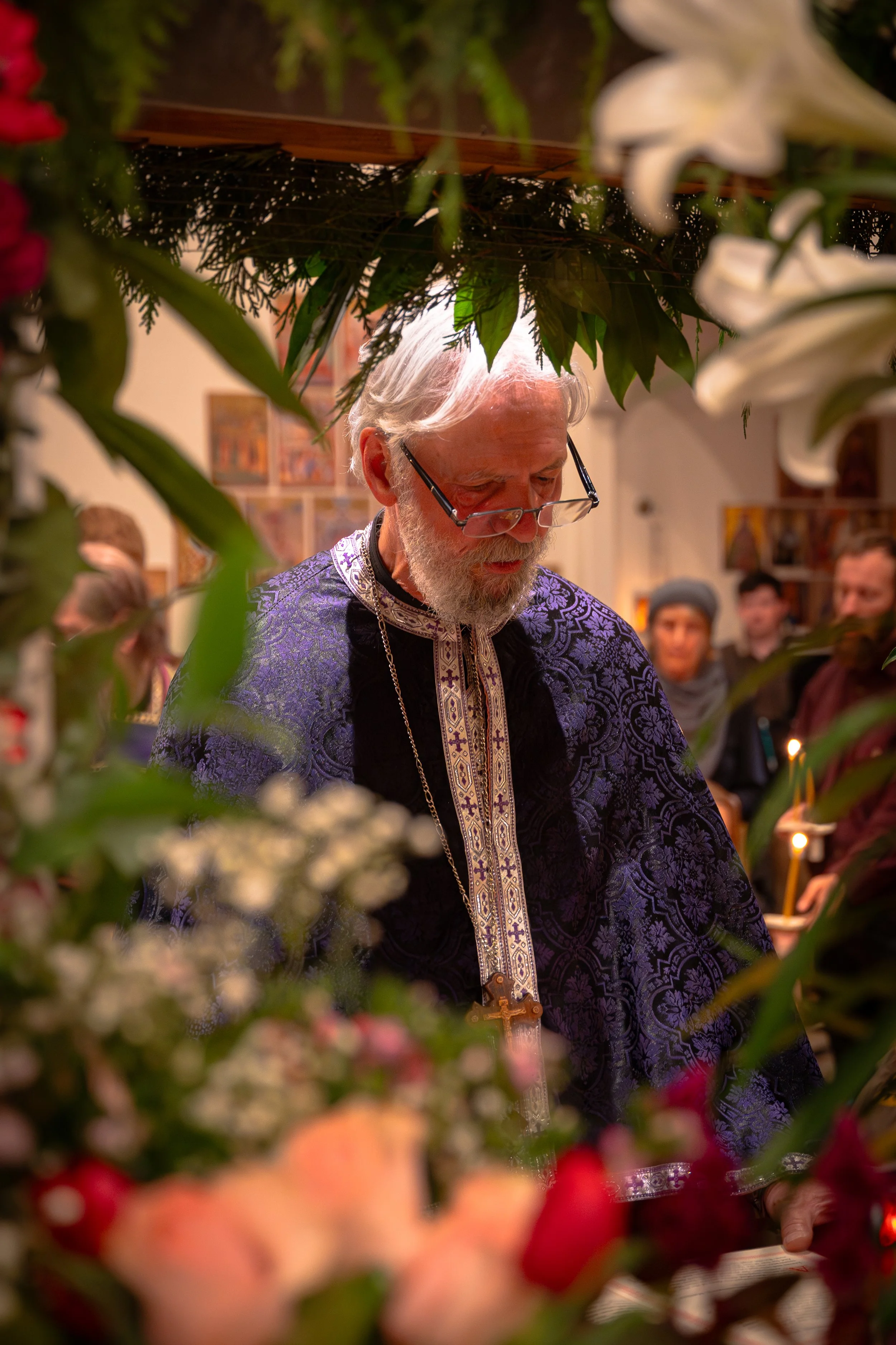 An elderly man with white hair, beard, and glasses, wearing a purple patterned robe with a cross on a chain around his neck, is seen inside a religious setting surrounded by flowers and greenery, likely participating in a religious ceremony.