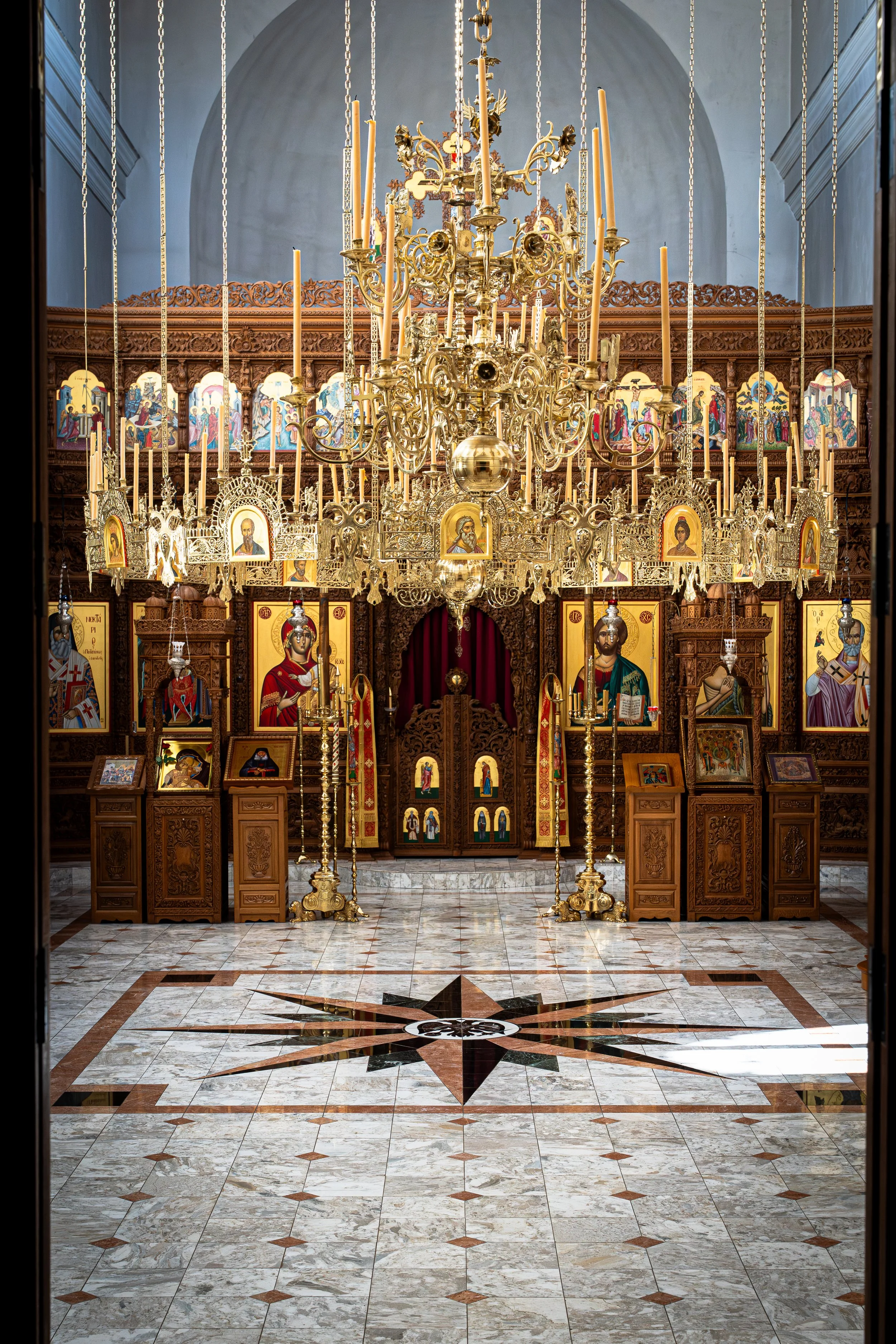 Interior view of an ornate church or cathedral, showcasing a large, intricate gold chandelier hanging in front of religious icons and wood carvings, with a marble floor featuring a star pattern.
