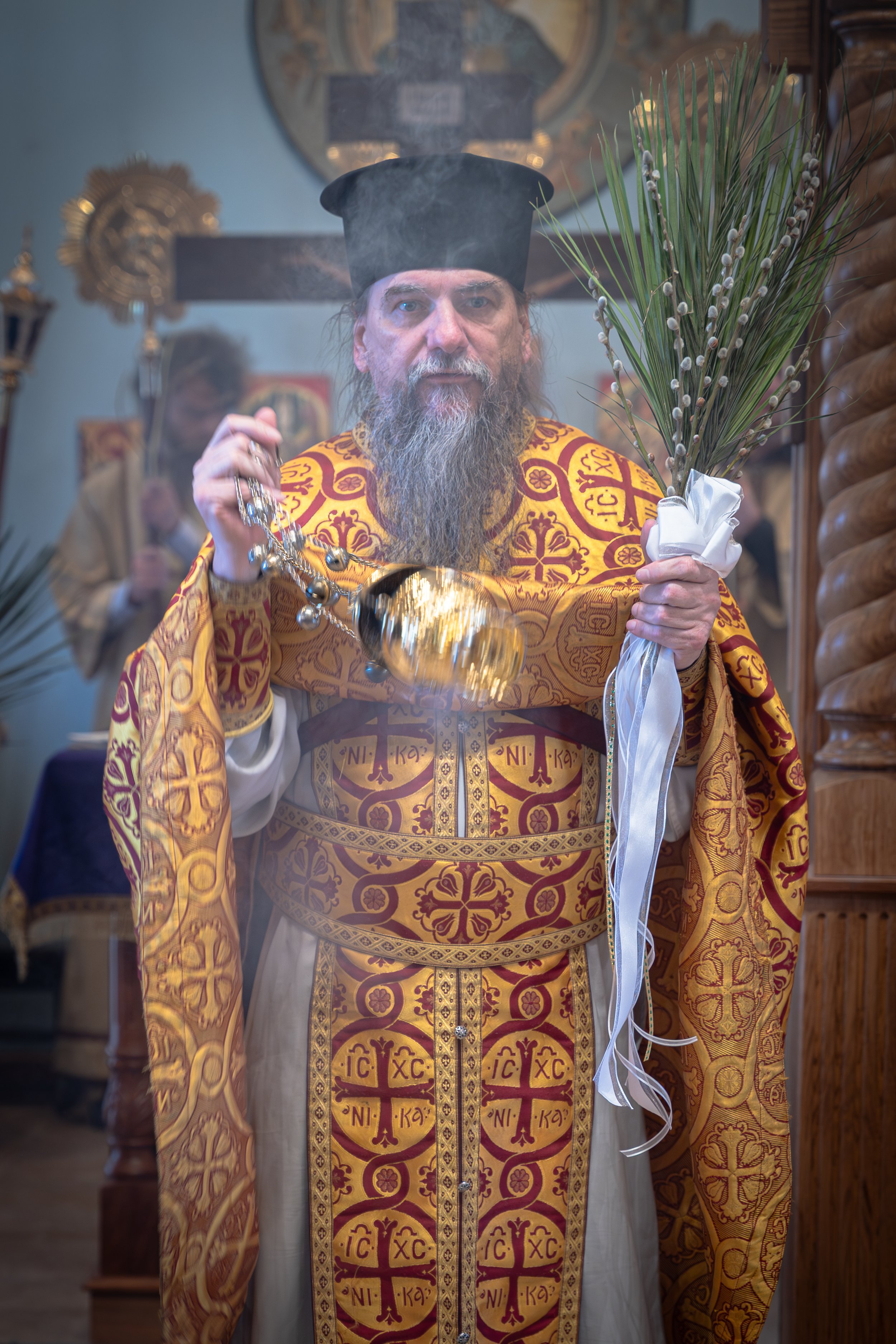 A priest in ornate gold and red vestments holding a bouquet of green branches with white blossoms, standing in a church with icons and religious artifacts visible in the background.