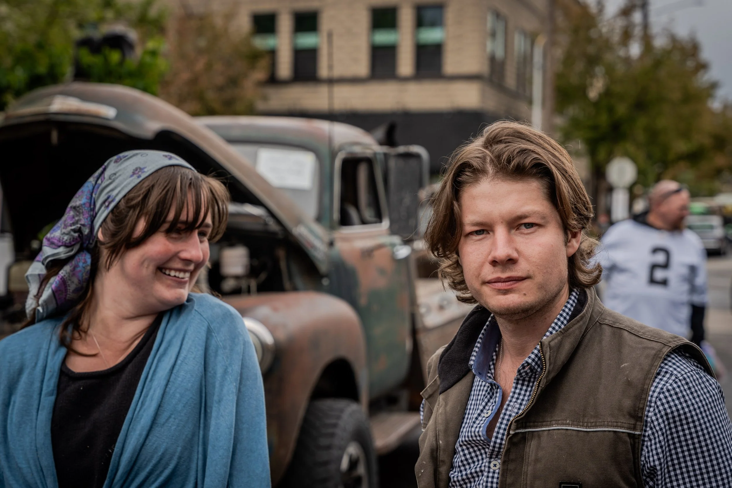 People standing outdoors in front of a vintage vehicle with a building and trees in the background.