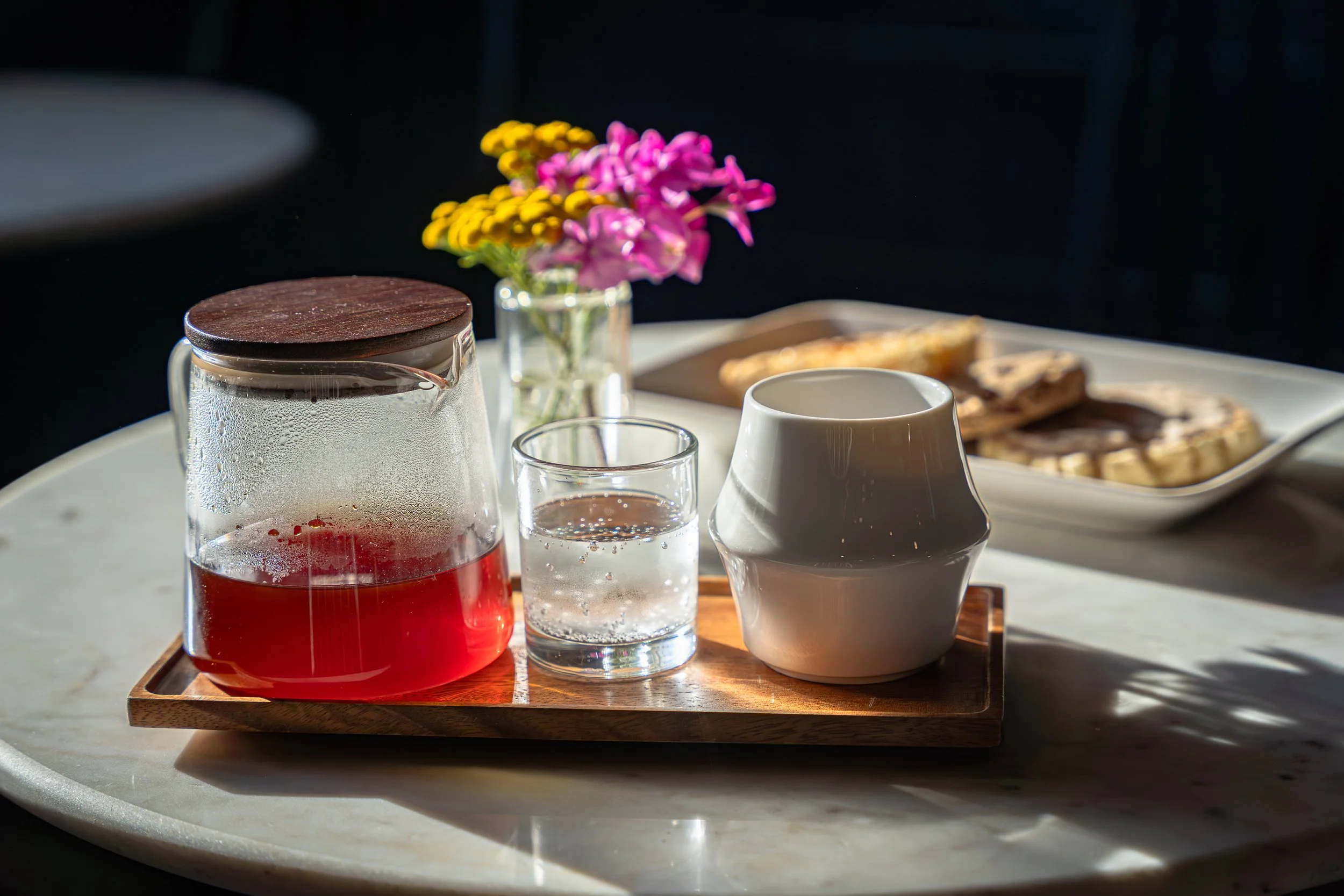 A wooden tray holding a glass jar of red juice with condensation, a small glass of clear water, and a white ceramic pitcher, with a vase of pink and yellow flowers and some snacks in the background on a tray.